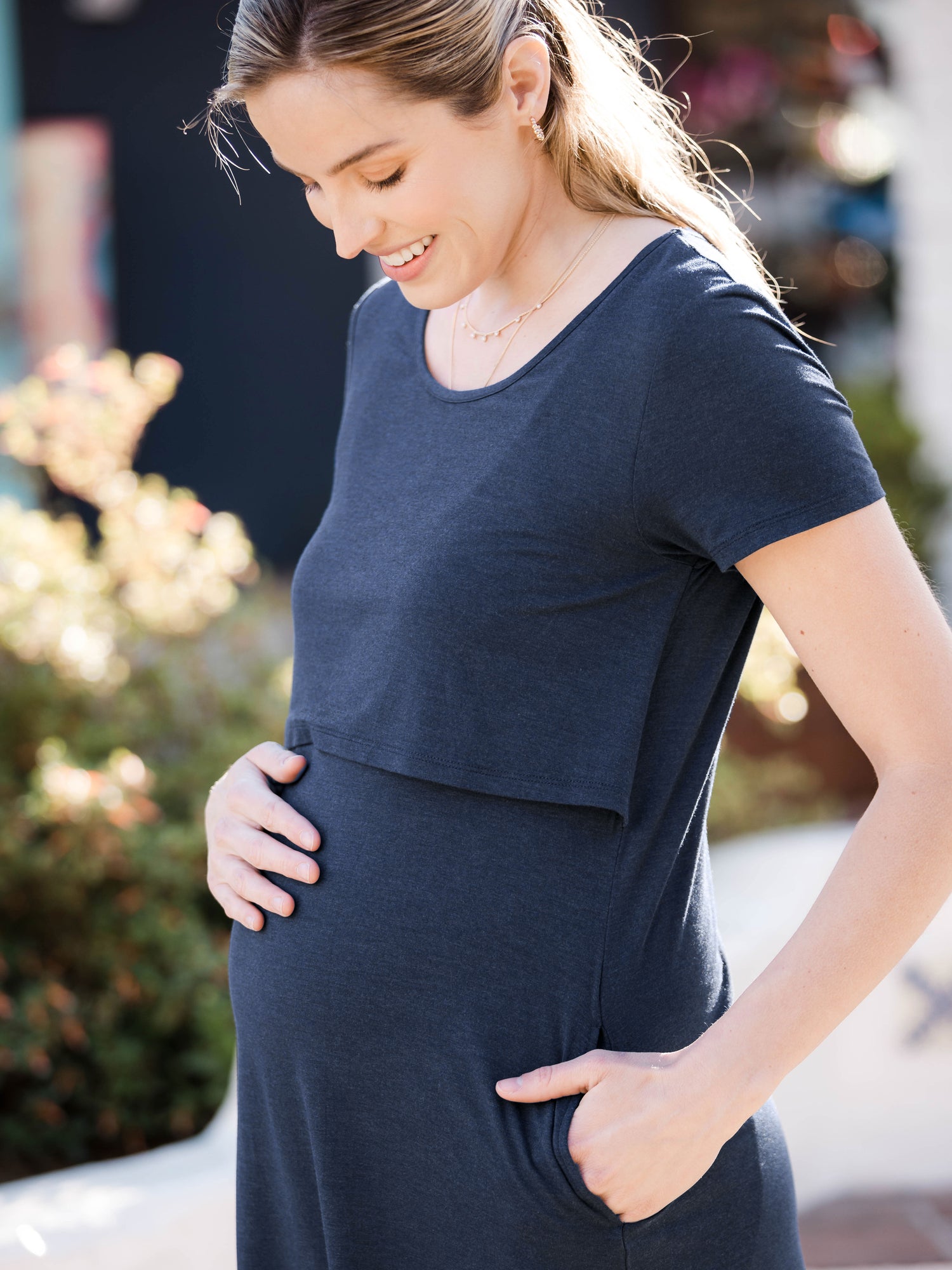 Closeup of a model outside wearing the  Eleanora Bamboo Maternity & Nursing Dress in Navy Heather and holding her baby bump.