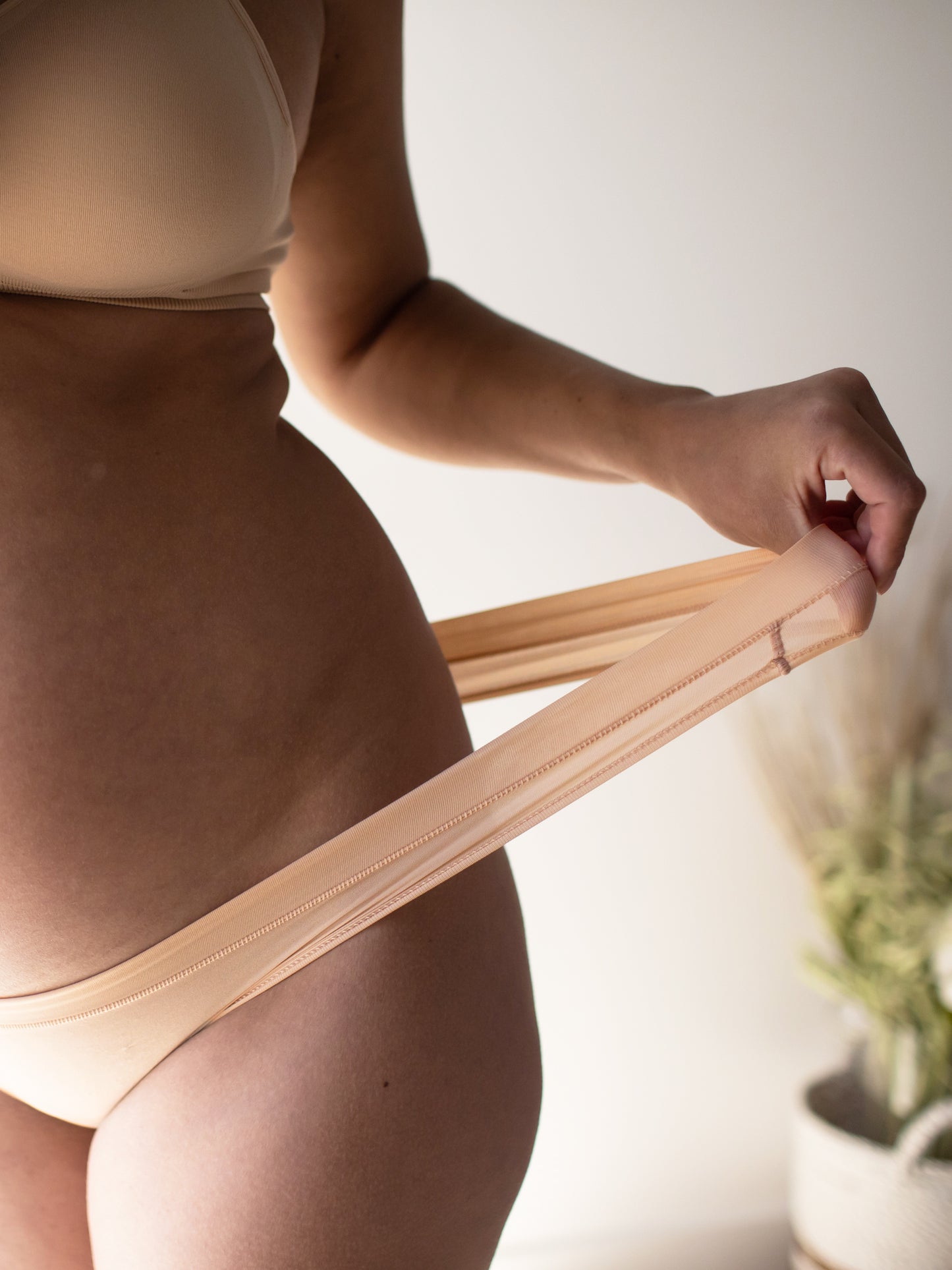 Closeup of a pregnant model tugging on the waistband of the Grow with Me™ Maternity & Postpartum Thong in Beige