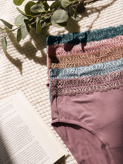 Product image of the High Waisted Postpartum Underwear Pack in Dusty Hues against a woven beige mat with eucalyptus leaves and a book