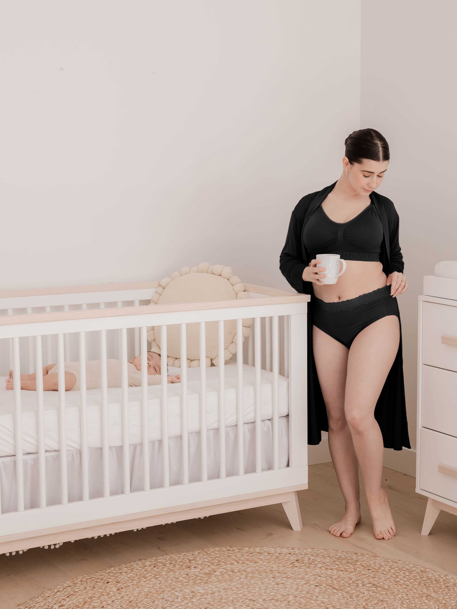 Model standing next to a crib holding her sleeping baby while wearing the  High-Waisted Postpartum Underwear in Assorted Neutrals. @model_info:Kianna is wearing a Small.
