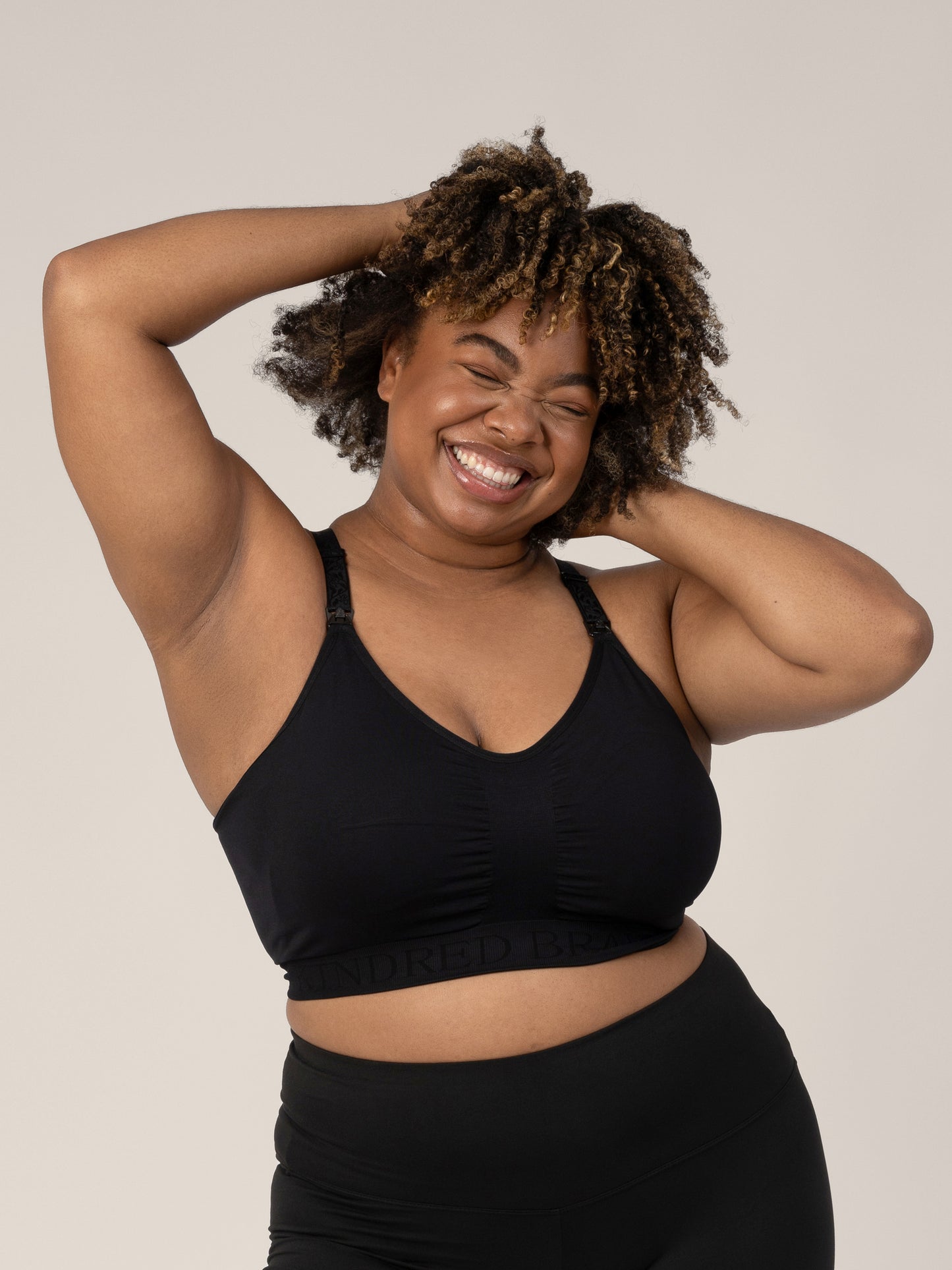 Model wearing the Sublime® Nursing Sports Bra in Black with her hands in her hair and giving a big smile. @model_info: Skye is wearing an X-Large Busty.