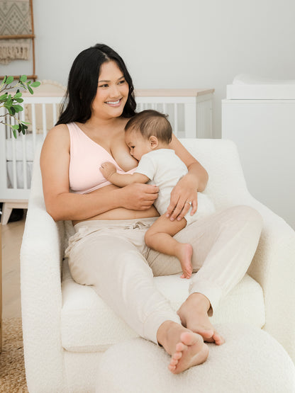 Model sitting in a chair with her baby breastfeeding while wearing the Sublime® Adjustable Crossover Nursing & Lounge Bra in Soft Pink
