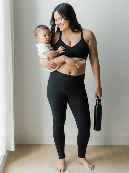 Model wearing the Sublime® Hands-Free Pumping & Nursing Sports Bra in Black with her baby on her hip holding a metal water bottle.