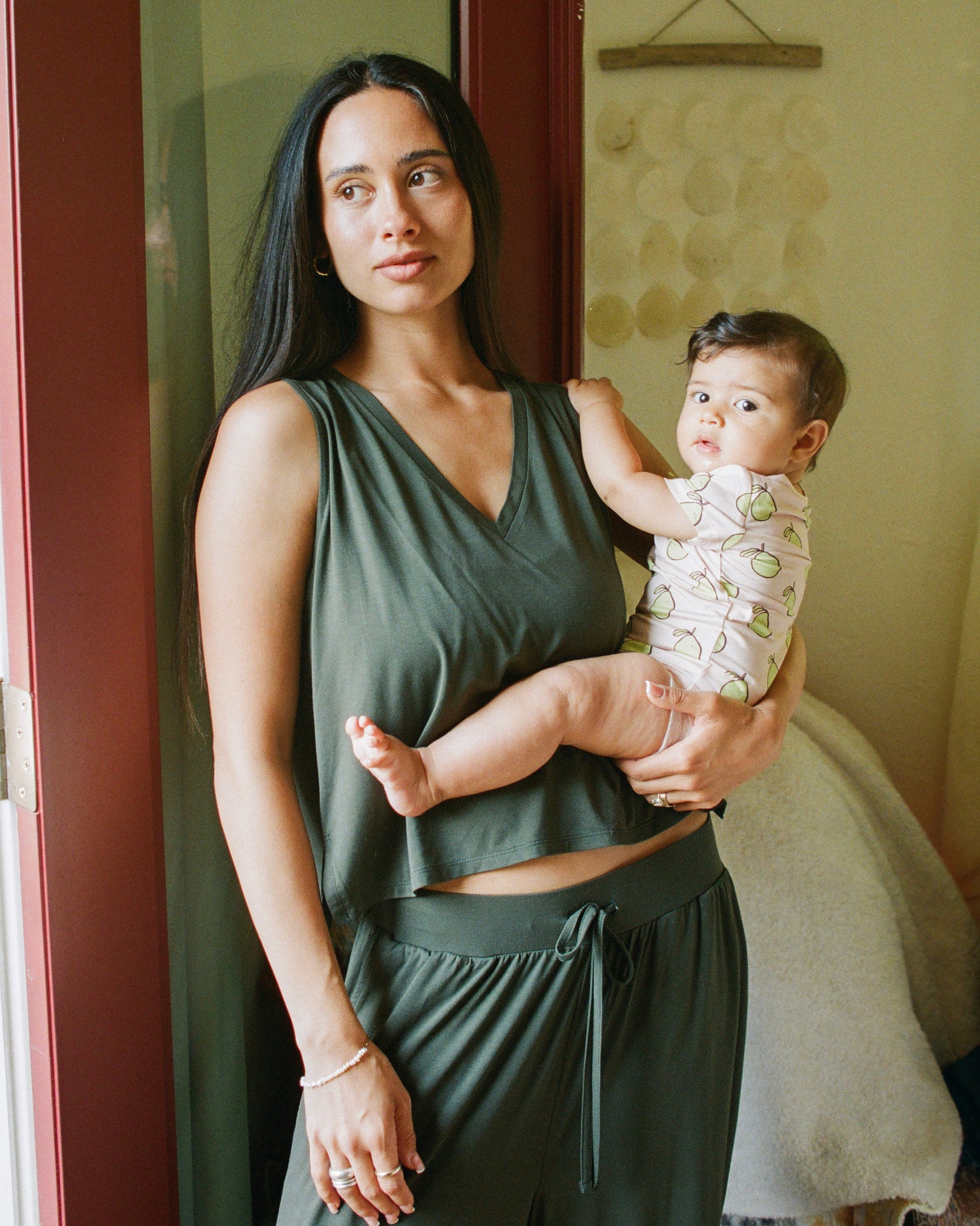 Model is holding baby wearing Onesie in Pink Pomelo, and model is wearing Aria Bamboo Cropped V-neck Tank in Thyme and Aria Bamboo Casual Lounge Pant in Thyme in a domestic front entrance.