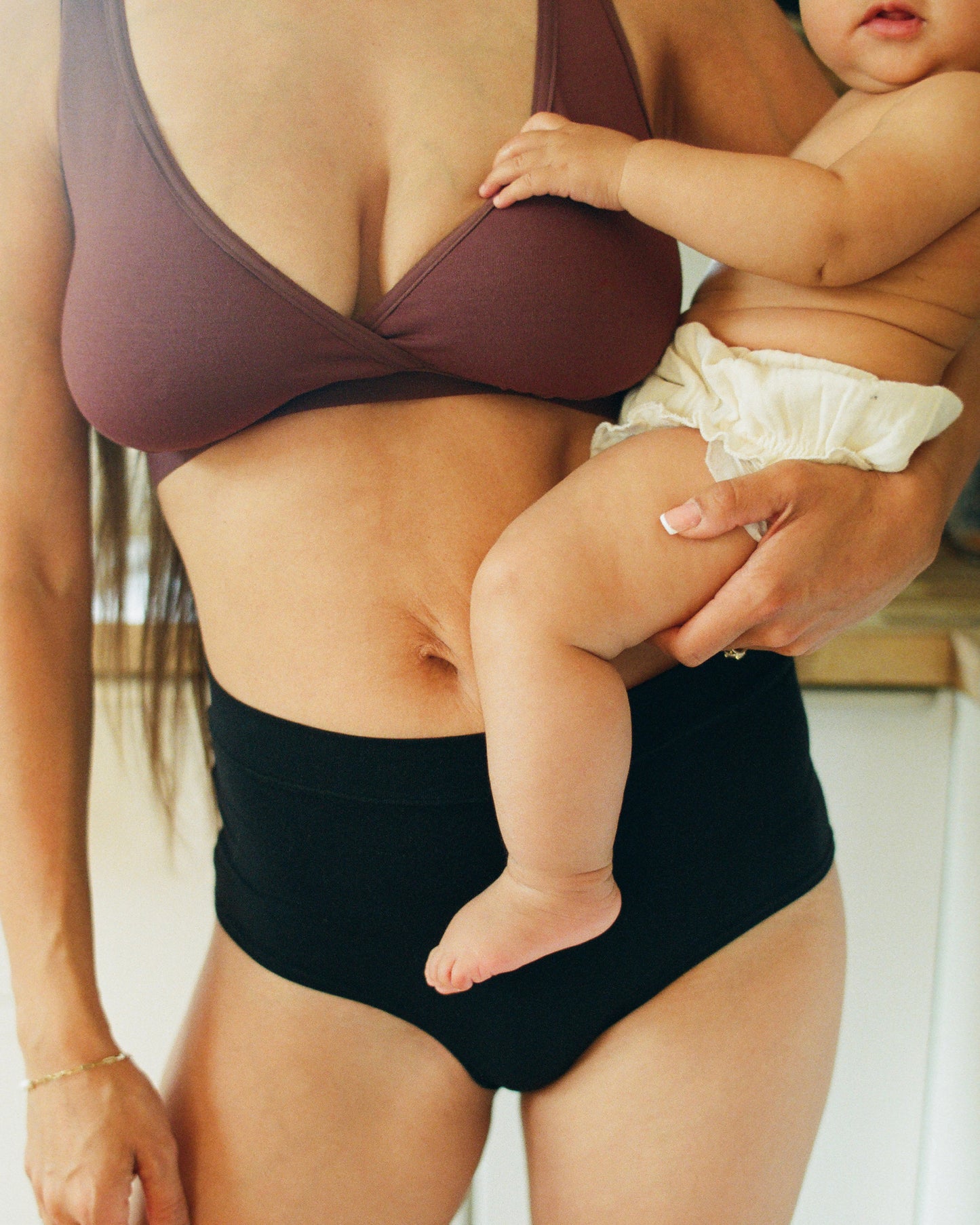 Model is holding baby and wearing Every Stage Crossover Bra in Cocoa and Every Stage Brief in Black with a kitchen background