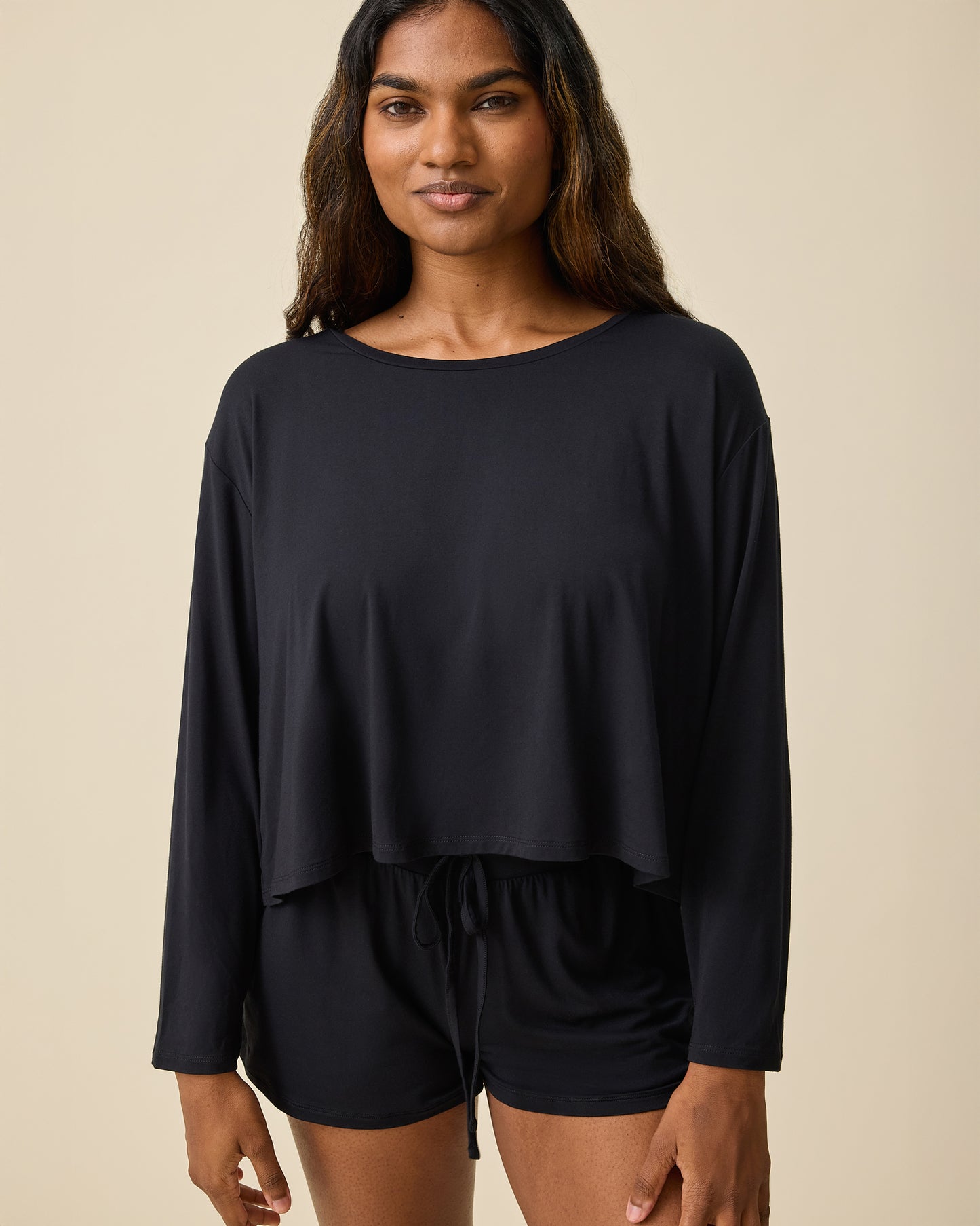 Woman wearing the Aria Bamboo Casual Cropped Long Sleeve Top in black with matching shorts on a beige background@model_info:Sahana is wearing a Small.