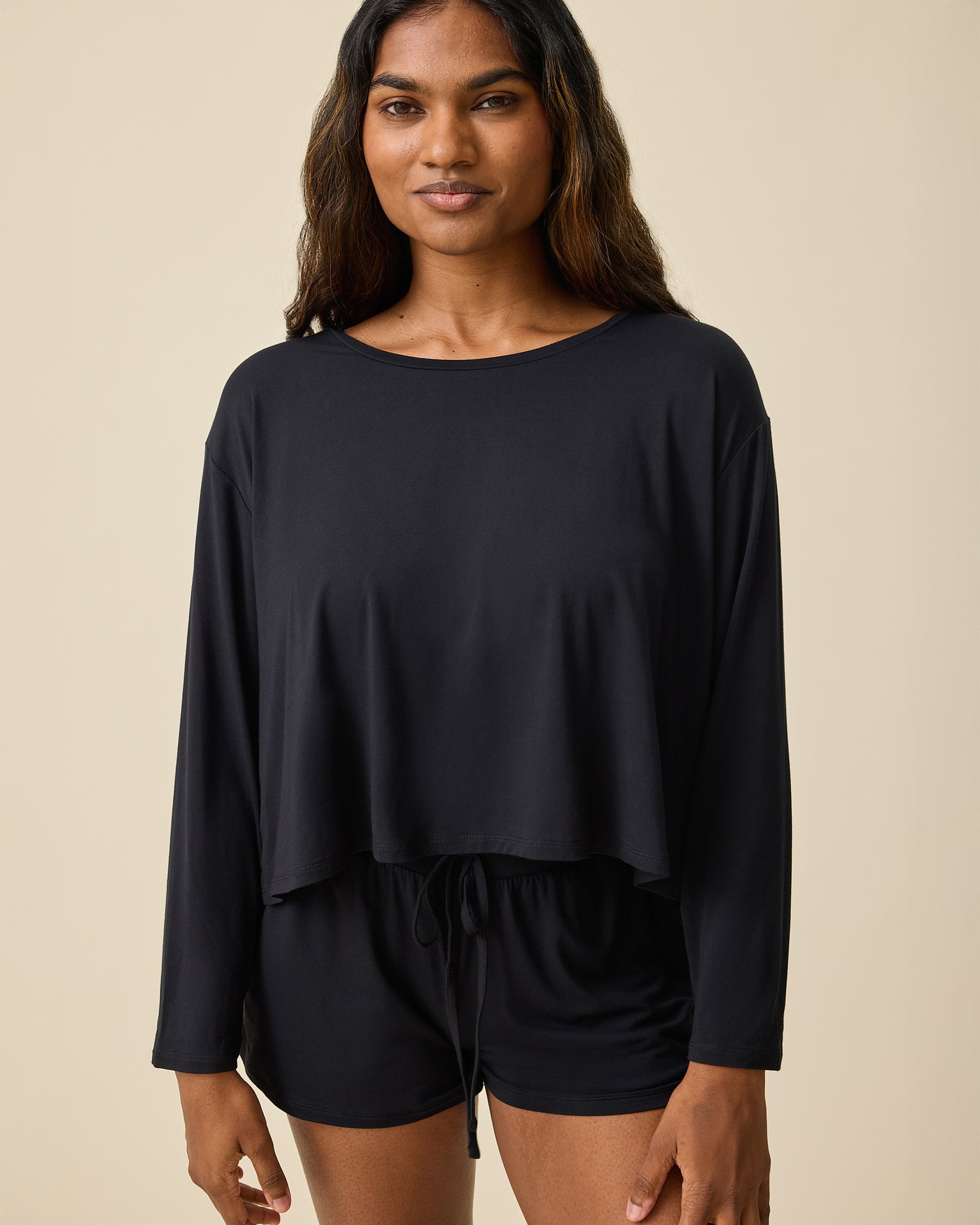 Woman wearing the Aria Bamboo Casual Cropped Long Sleeve Top in black with matching shorts on a beige background@model_info:Sahana is wearing a Small.