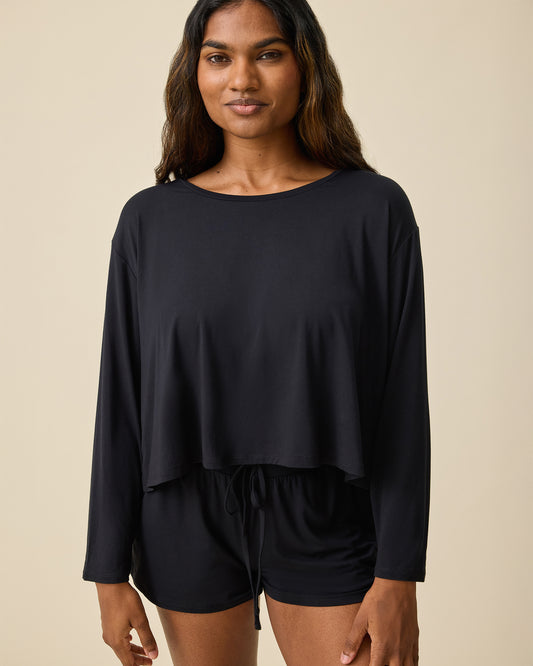 Woman wearing the Aria Bamboo Casual Cropped Long Sleeve Top in black with matching shorts on a beige background@model_info:Sahana is wearing a Small.
