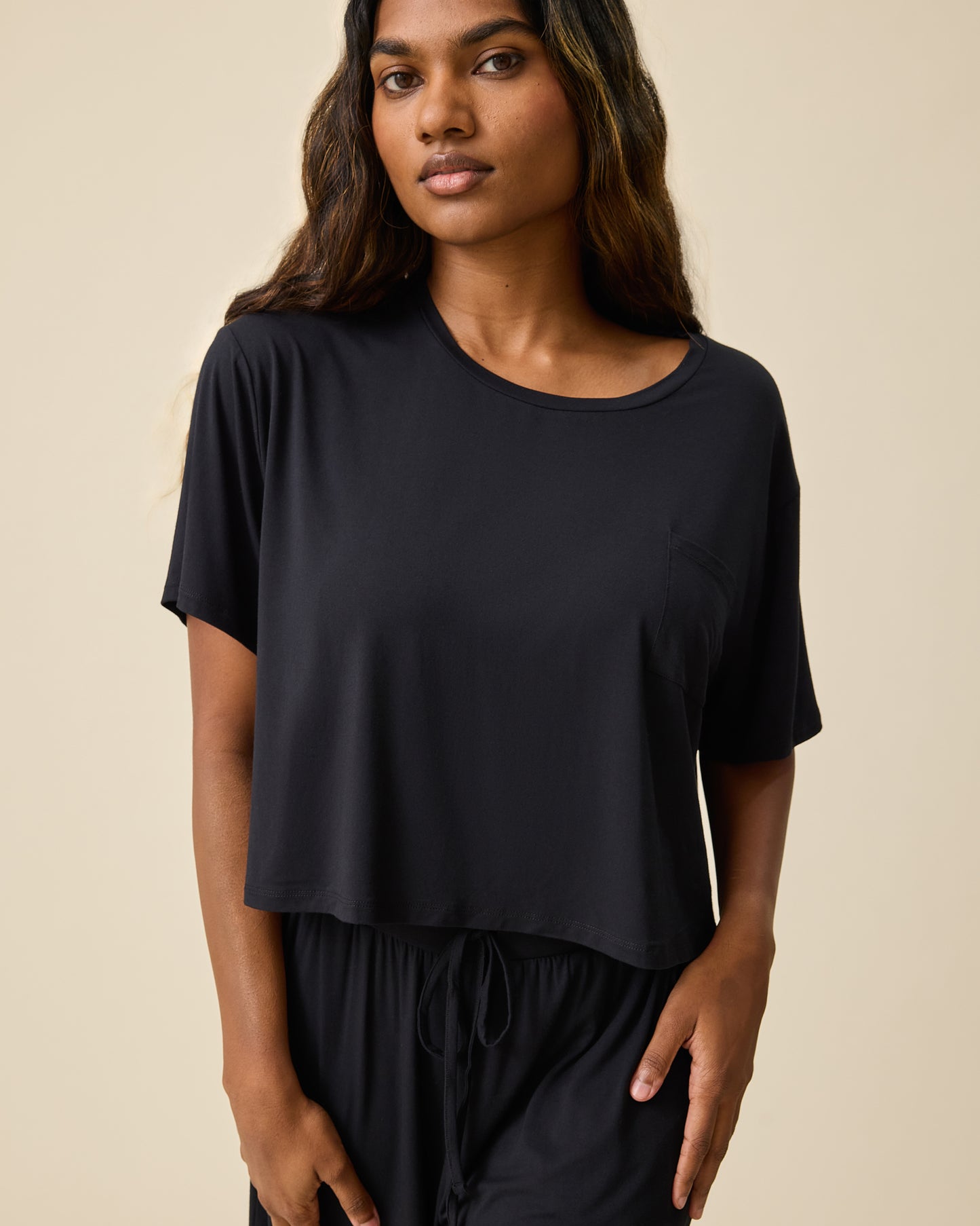 Woman wearing the Aria Bamboo Casual Cropped Short Sleeve Top in black with matching pants on beige background@model_info:Sahana is wearing a Small.