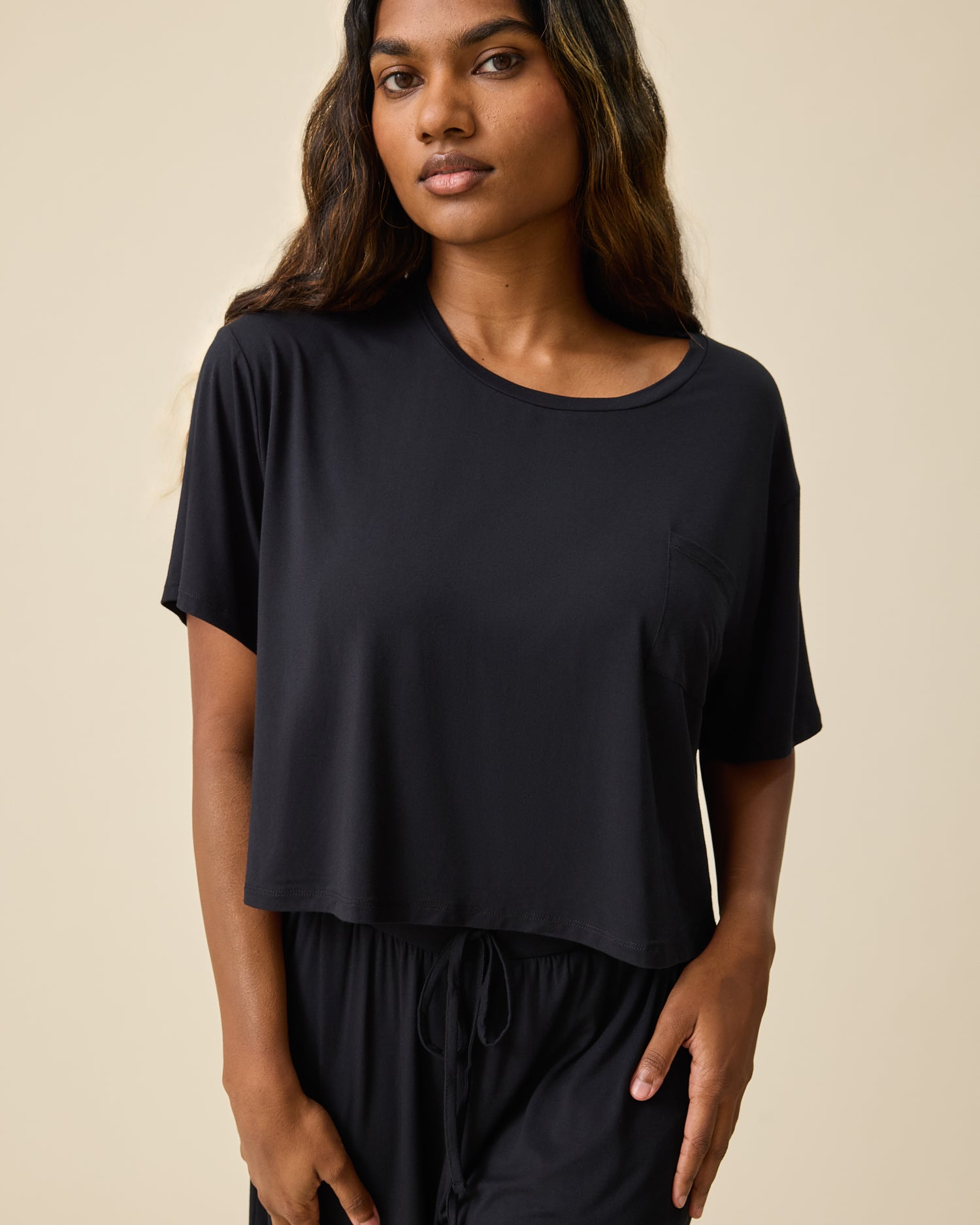 Woman wearing the Aria Bamboo Casual Cropped Short Sleeve Top in black with matching pants on beige background@model_info:Sahana is wearing a Small.