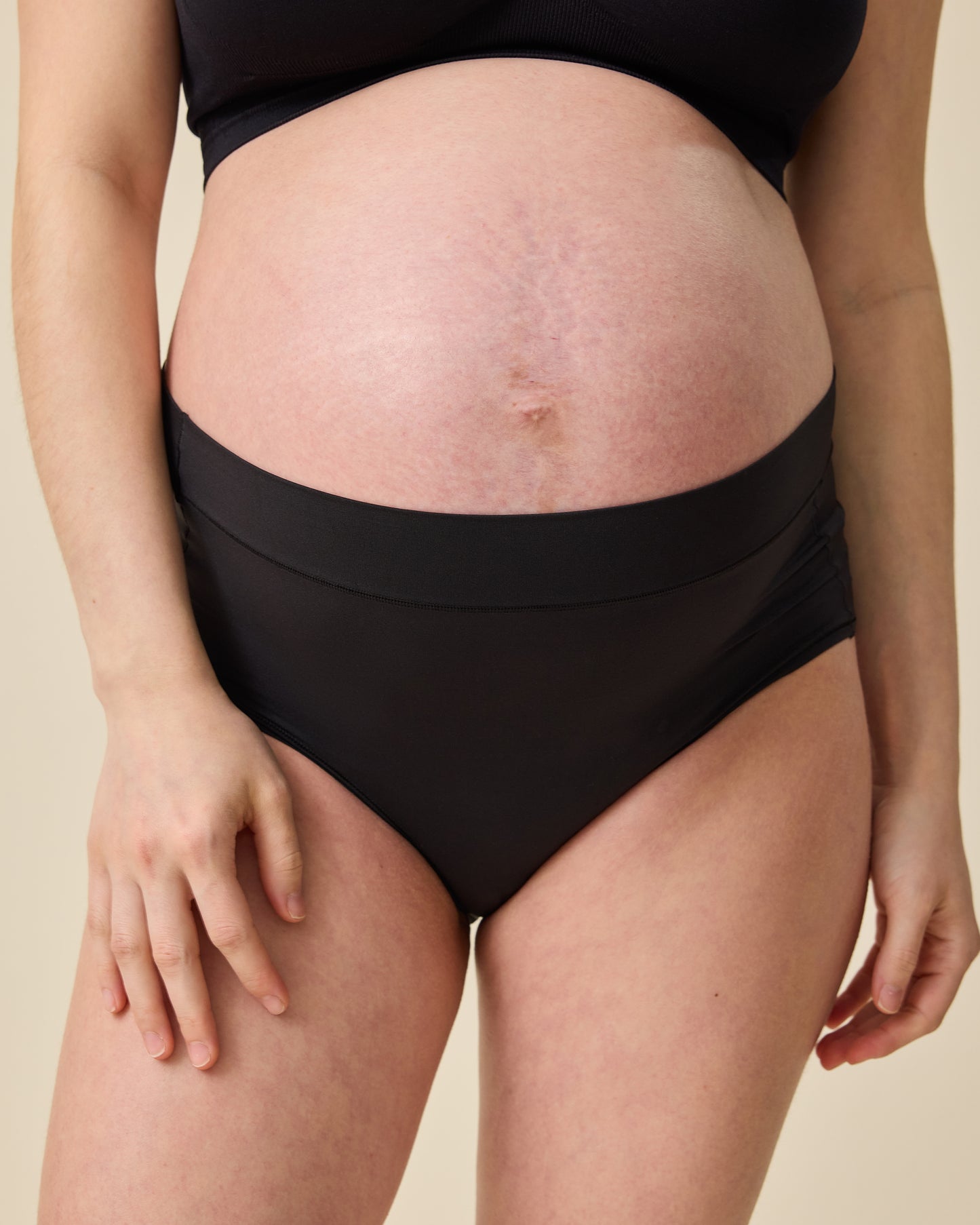 Close up of model wearing Grow with Me™ Maternity & Postpartum Brief in Black on a beige background@model_info:Tassia is wearing a Medium.