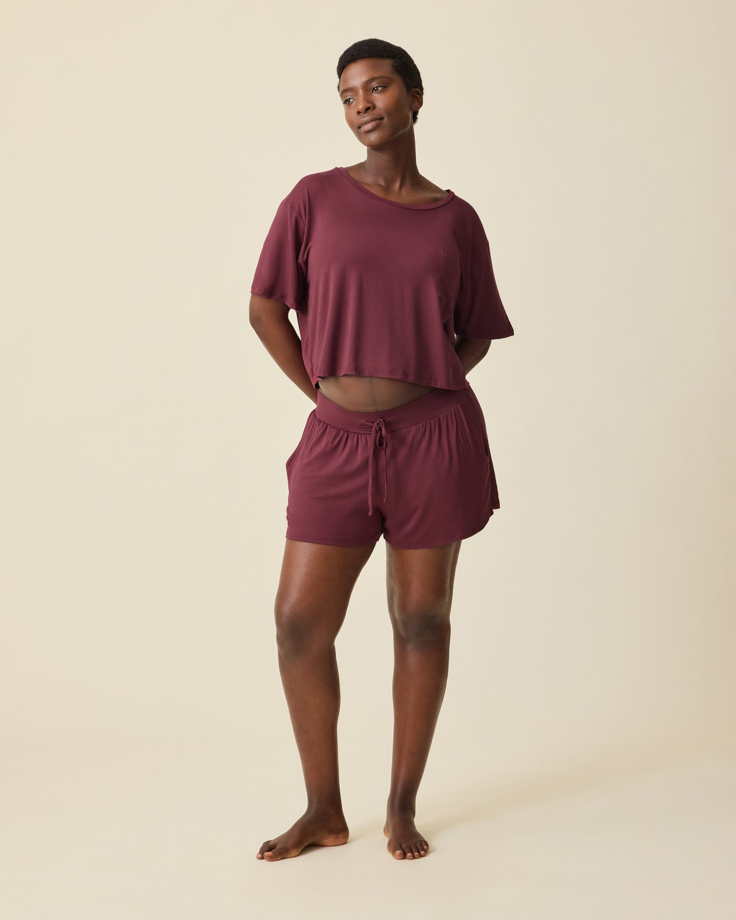 Pregnant model is wearing Aria Bamboo Casual Cropped Short Sleeve Top in Deep Burgundy and Aria Bamboo Casual Lounge Short in Deep Burgundy, on a beige background. @model_info:Anna is 5'8.5" and wearing a Medium.