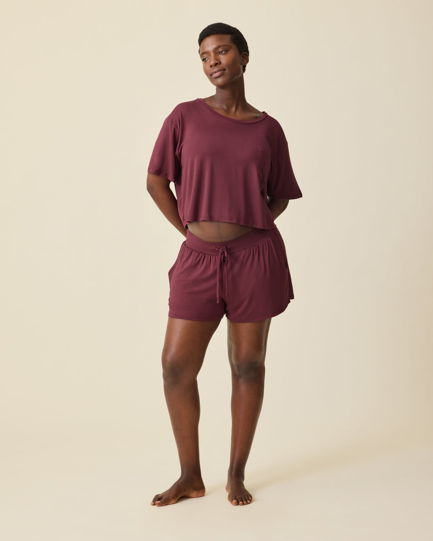 Pregnant model is wearing Aria Bamboo Casual Cropped Short Sleeve Top in Deep Burgundy and Aria Bamboo Casual Lounge Short in Deep Burgundy, on a beige background. @model_info:Anna is 5'8.5" and wearing a Medium.