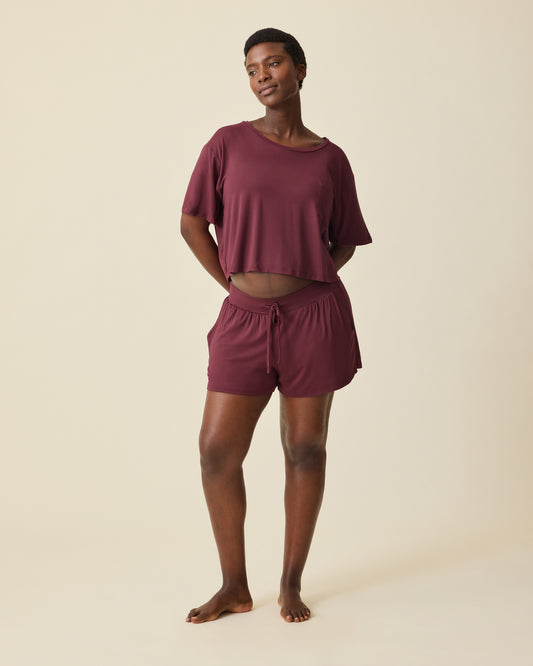 Pregnant model is wearing Aria Bamboo Casual Cropped Short Sleeve Top in Deep Burgundy and Aria Bamboo Casual Lounge Short in Deep Burgundy, on a beige background. @model_info:Anna is 5'8.5" and wearing a Medium.