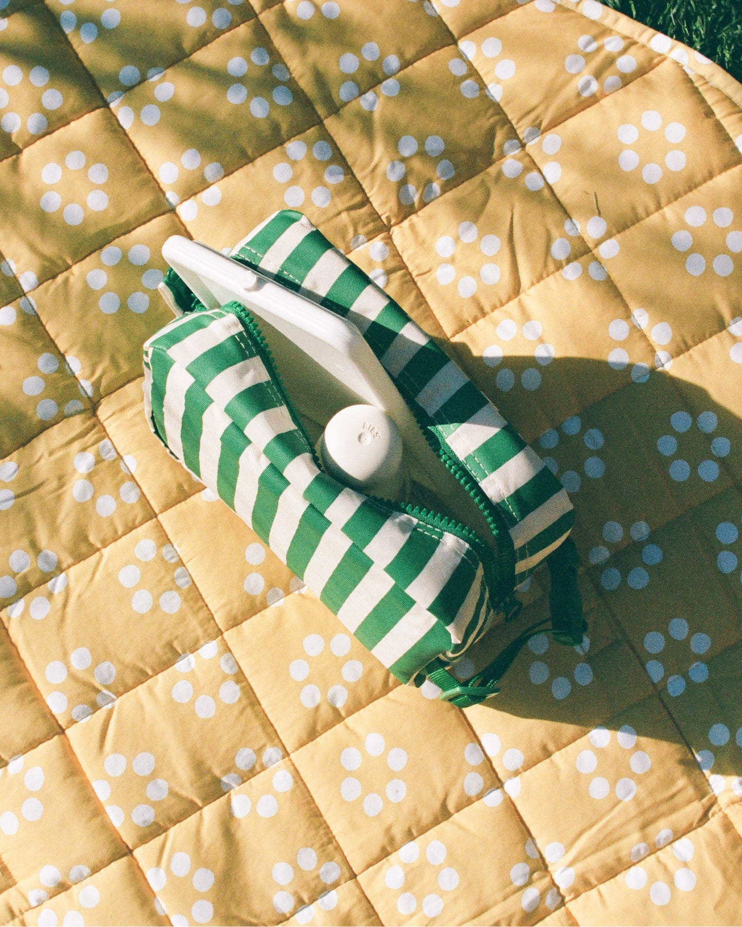 Mini Cooler Bag in Green Stripe holding bottle and wipes container, sitting on an Inside/Outside Play Mat in Sunshine Dots, on a grassy background.