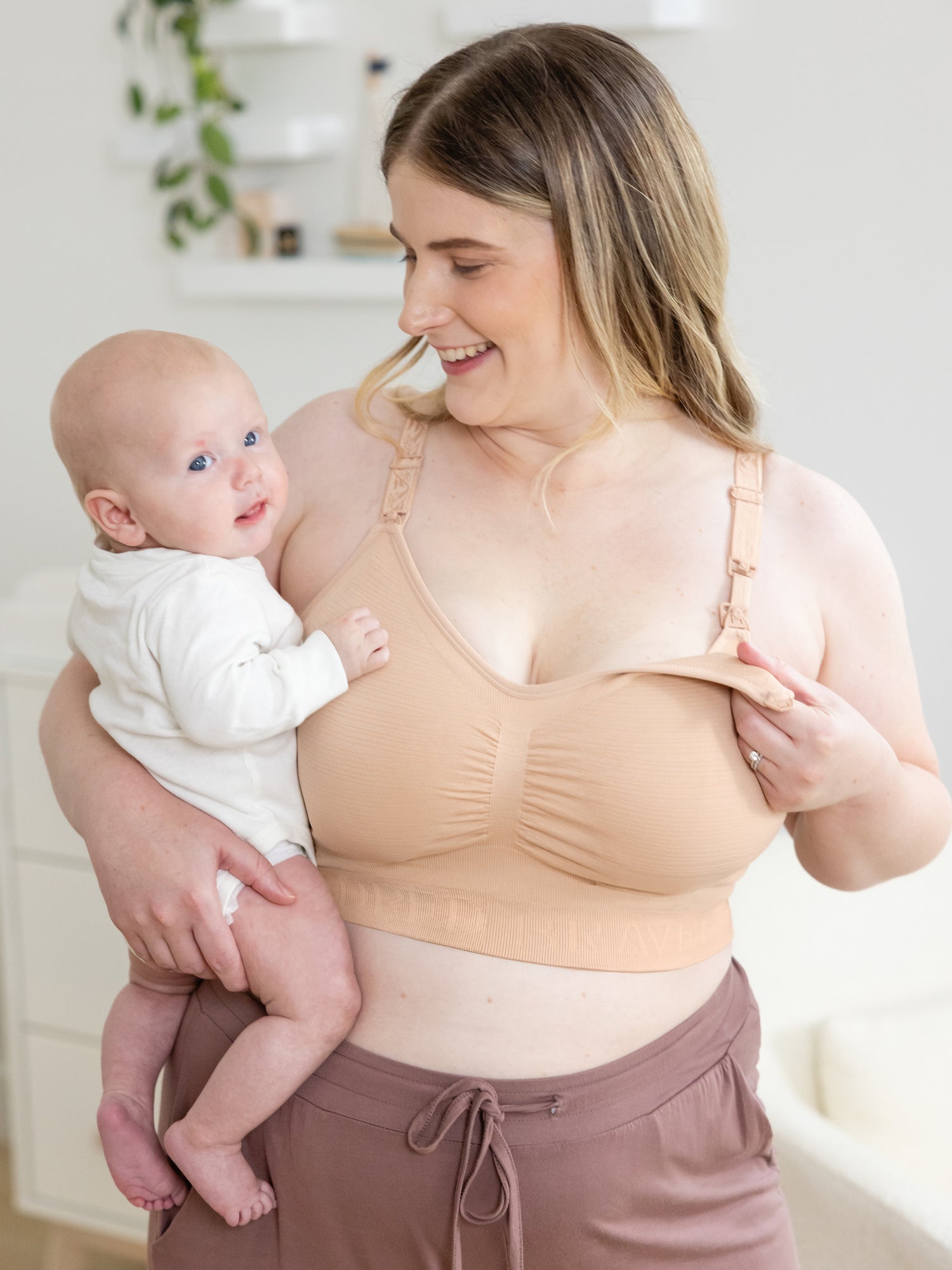 Model holding baby in one arm and showing unclipping feature on the  Sublime® Hands-Free Pumping & Nursing Bra in beige. @model_info:Piper is wearing a Large Super Busty.