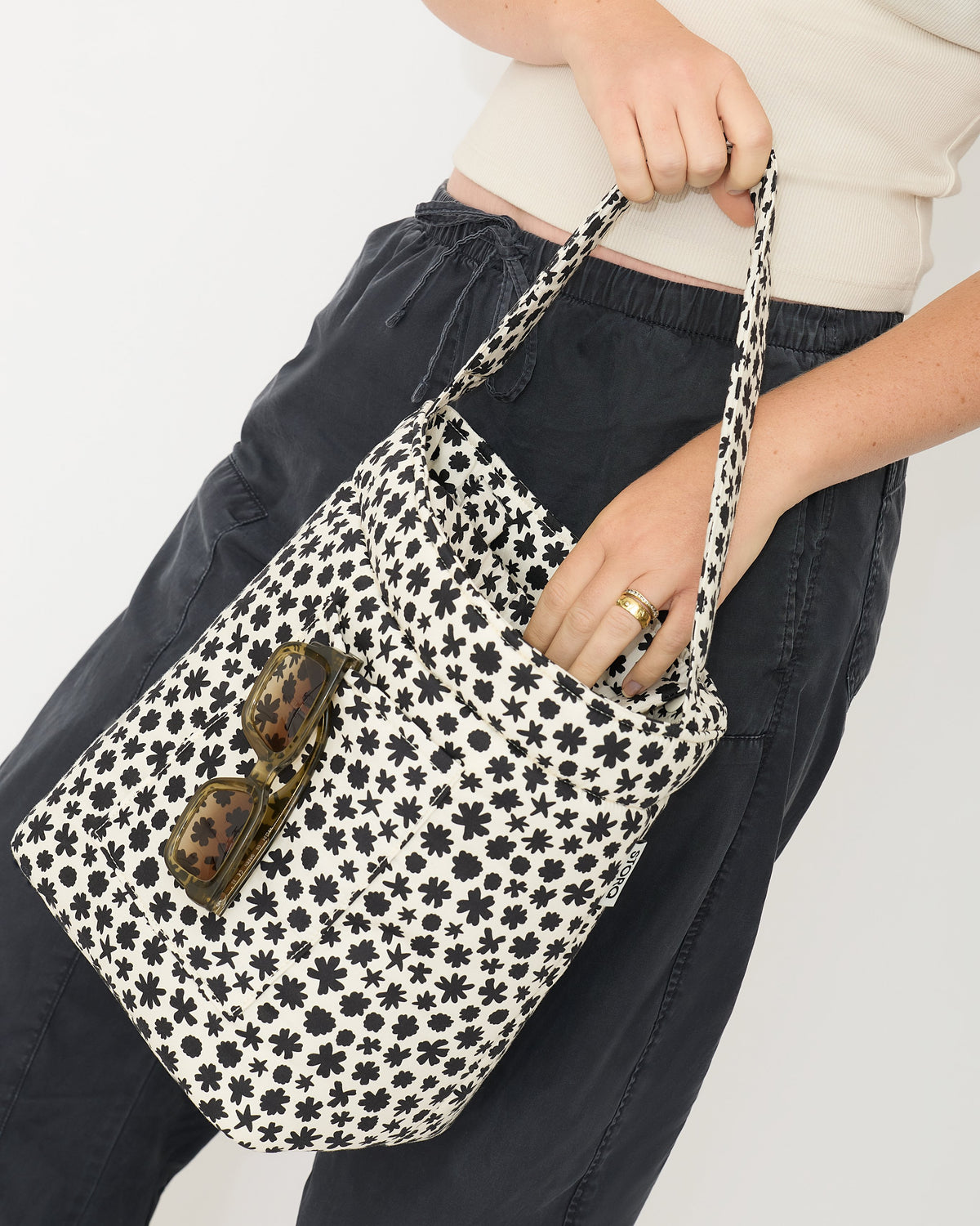Model is holding Stroller Party™ Bucket Bag in Cream Chamomiles by the long strap, and reaching into the bag. Shades are hanging out of outer pocket. On a beige background.