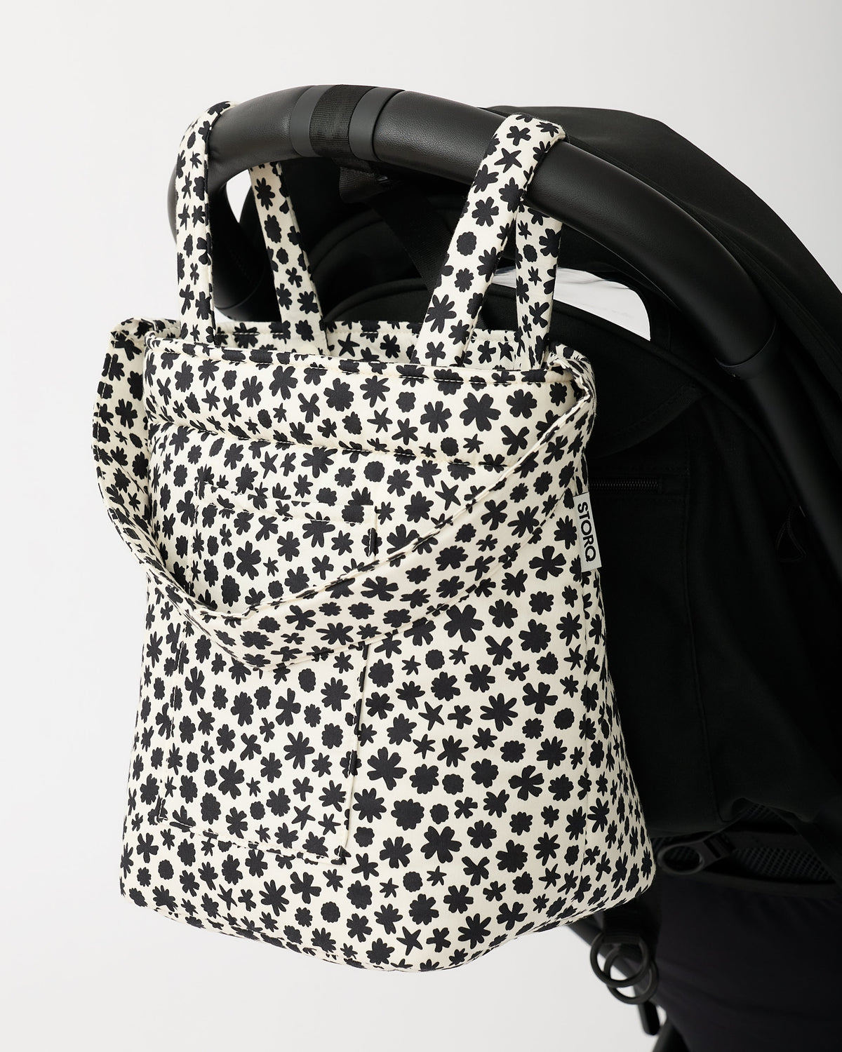 Stroller Party™ Bucket Bag in Cream Chamomiles is hanging off the handlebar of a black stroller, on a beige background.