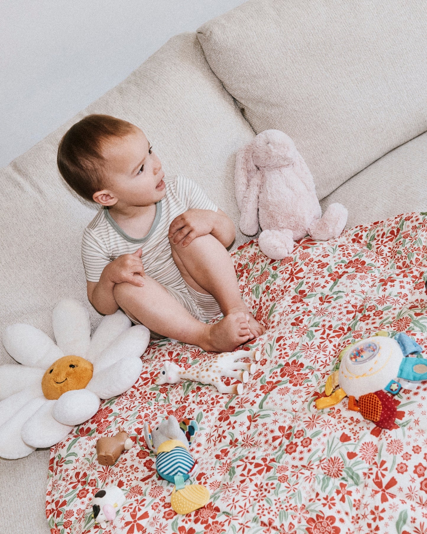 Toddler sitting on Inside/Outside Play Mat in Superbloom on a sofa with daisy pillow, bunny doll, Sophie the giraffe and various other tactile toys, on a white wall background.