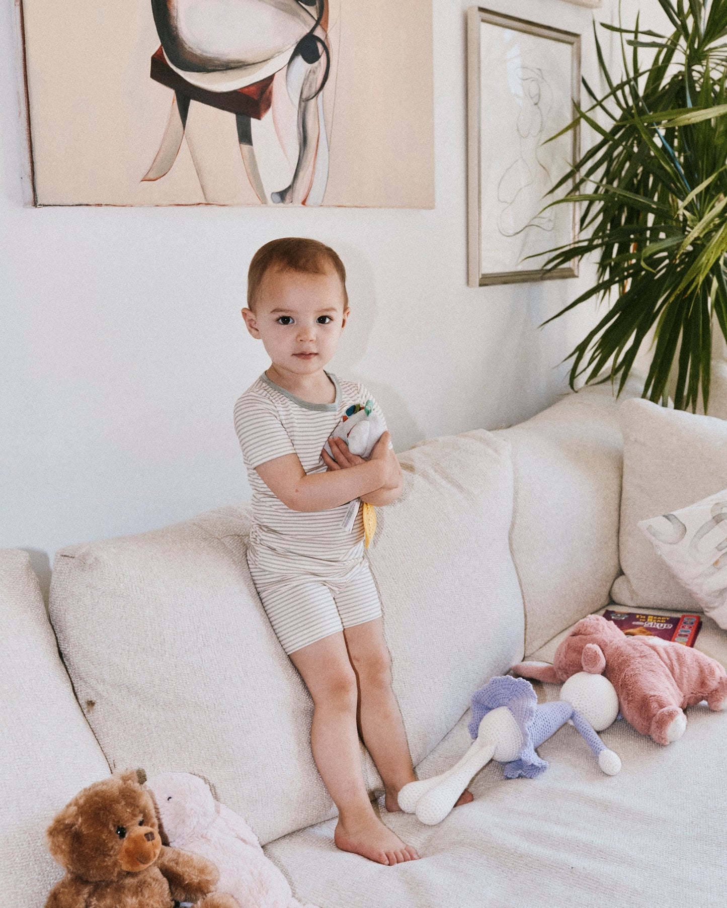 Toddler standing on white sofa, wearing Bamboo Short Sleeve & Shorts Set in Sage Candy Stripe and holding doll, amongst other dolls on sofa. Art and exotic plants in background.@model_info:Emi is wearing 18-24 months.