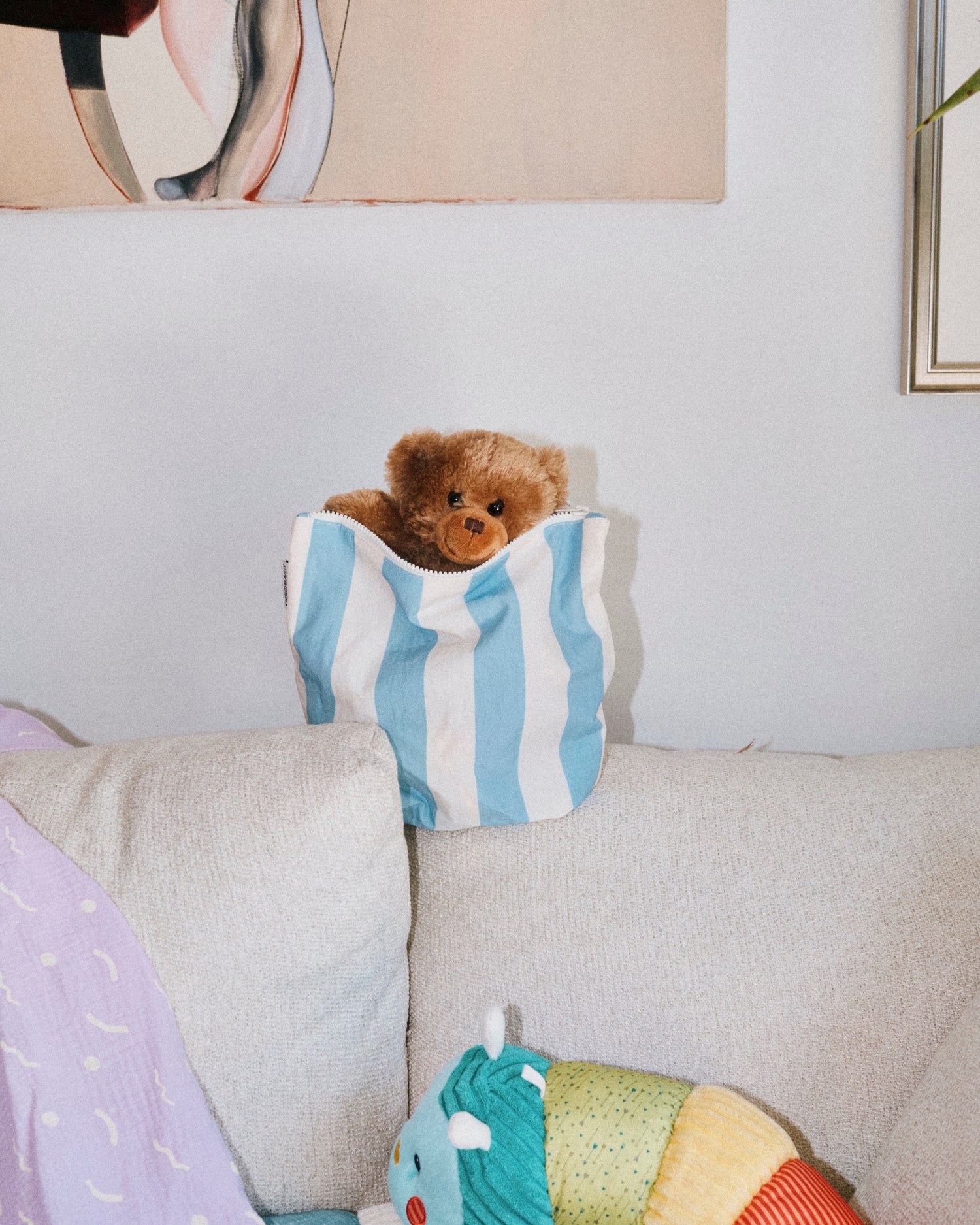Teddy bear peeking out of Large bag of the Zipper Pouch Three-Piece Set in Stripe, on a white sofa, next to caterpillar doll and Organic Muslin Swaddle in Lilac Squiggles, on a white wall background