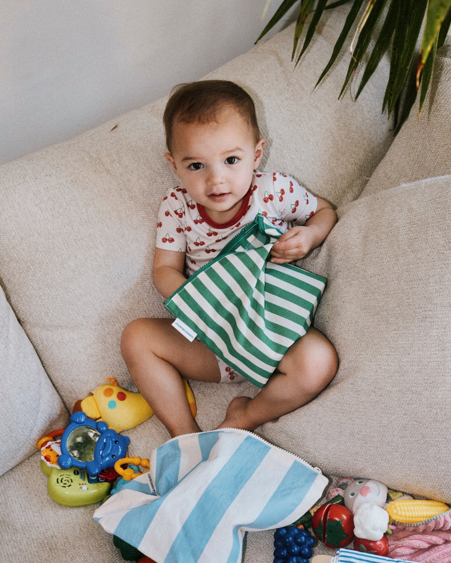 Toddler wearing Bamboo Short Sleeve & Shorts Set in Ivory Cherries, sitting on couch with many toys, while reaching into Zipper Pouch Three-Piece Set in Stripe. 
