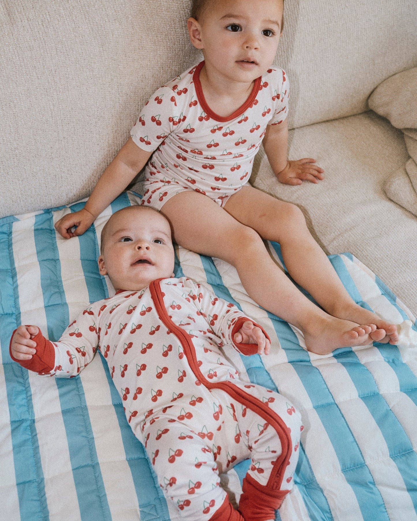 Toddler wearing Bamboo Short Sleeve & Shorts Set in Ivory Cherries, sitting on couch, covered by Inside/Outside Play Mat in Blue Wide Stripe with baby, wearing Bamboo Baby Pajama in Ivory Cherries.