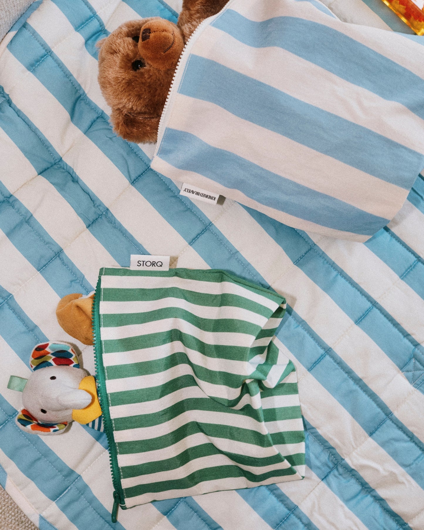 Zipper Pouch Three-Piece Set in Stripe, laying on the Inside/Outside Play Mat in Blue Wide Stripe. Each bag is holding dolls, toys and loveys.
