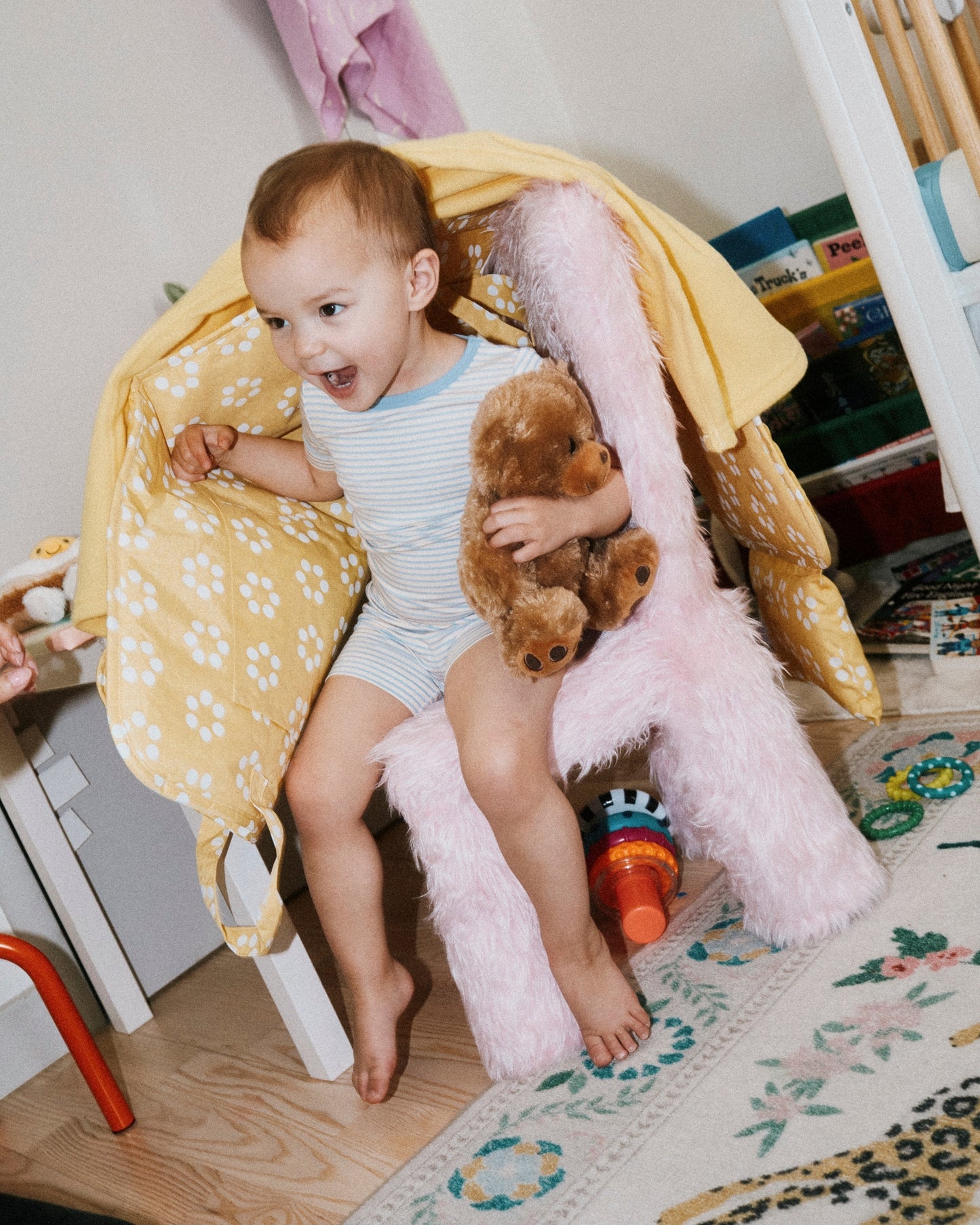 Toddler wearing Bamboo Short Sleeve & Shorts Set in Blue Candy Stripe, covered in Inside/Outside Play Mat in Sunshine Dots and holding teddy, sitting in a fuzzy pink chair, with playroom in background.