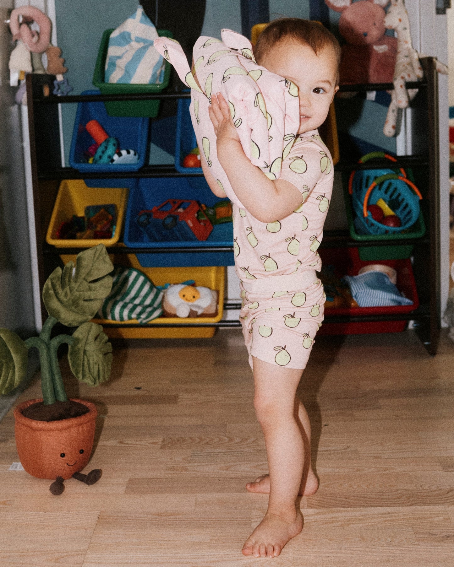 Toddler wearing Bamboo Short Sleeve & Shorts Set in Pink Pomelo and holding Inside/Outside Play Mat in Pink Pomelo in front of wall of toys in organizer. @model_info:Emi is wearing 18-24 months.