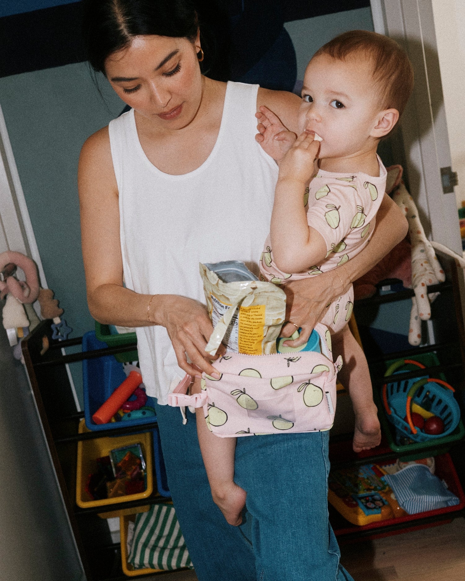 Woman holding toddler wearing Bamboo Short Sleeve & Shorts Set in Pink Pomelo while eating snacks from Mini Cooler Bag in Pink Pomelo. Woman is wearing Modal Signature Nursing Tank in White and Wide Leg Maternity & Postpartum Jean in Medium Wash, against a background of playroom full of toys and toy organizers.