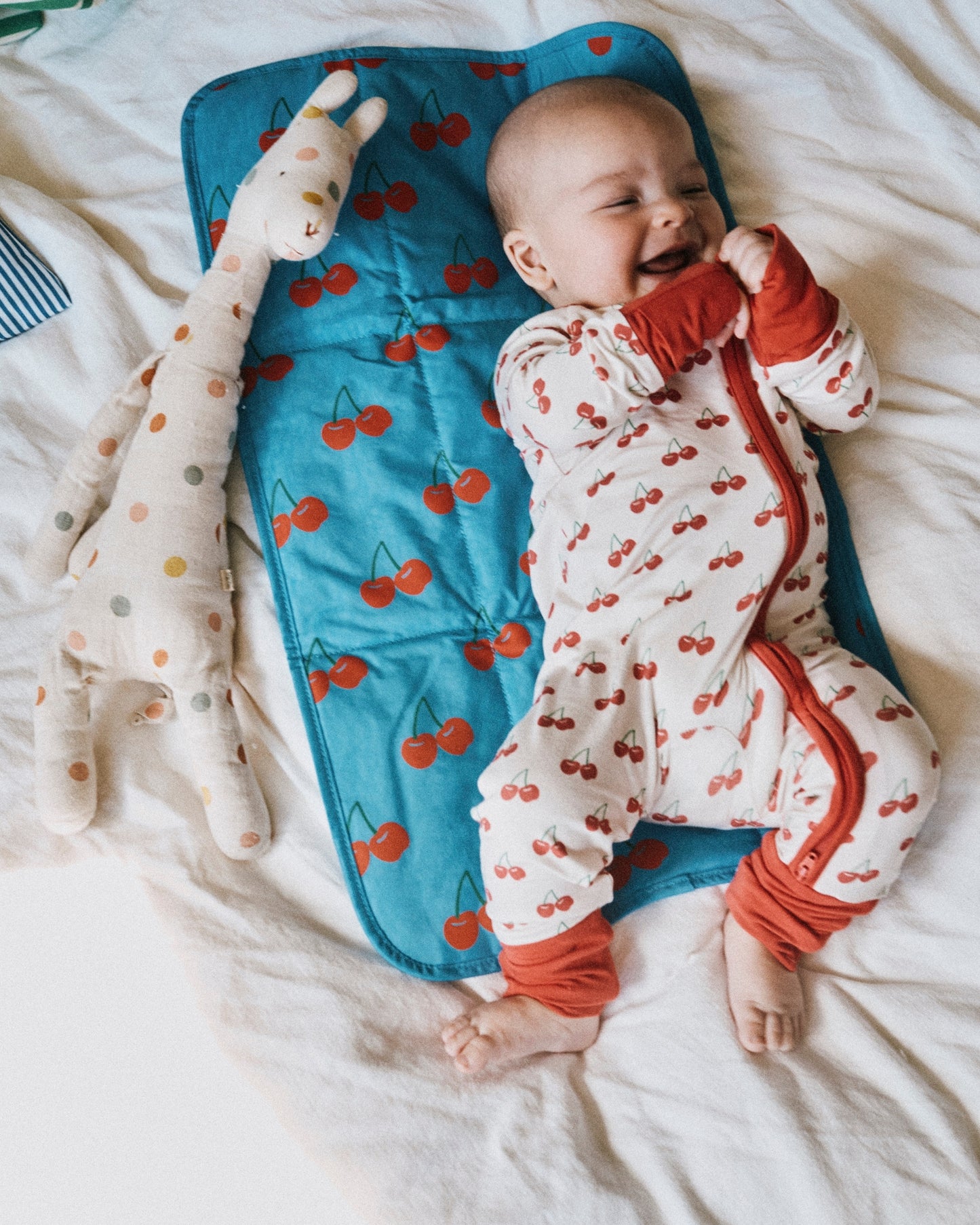 Smiling baby wearing Bamboo Baby Pajama | Ivory Cherries, laying on Quick Change Kit in Blue Cherries. Next to baby is Wet/Dry Bag Set in Stripe, doll and white bed background.@model_info:Johnny is wearing 3-6 months.