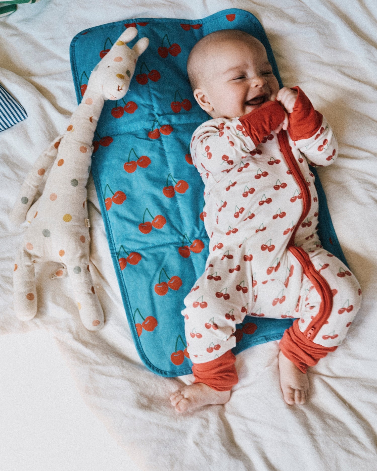 Smiling baby wearing Bamboo Baby Pajama | Ivory Cherries, laying on Quick Change Kit in Blue Cherries. Next to baby is Wet/Dry Bag Set in Stripe, doll and white bed background.@model_info:Johnny is wearing 3-6 months.
