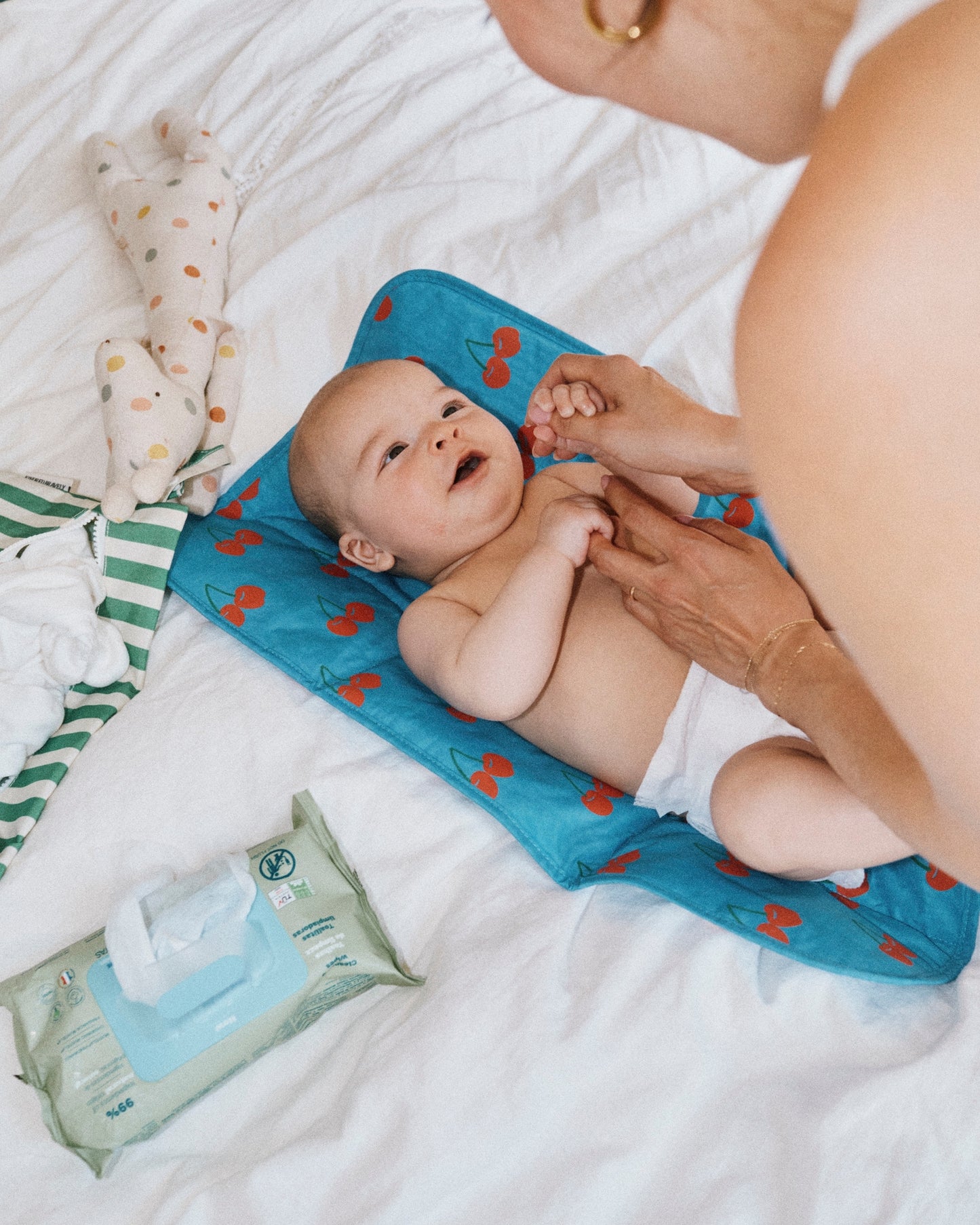 Woman lookin at smiling baby laying on Quick Change Kit in Blue Cherries. Next to baby is Wet/Dry Bag Set in Stripe, wet wipes, diapers, doll and white bed background.