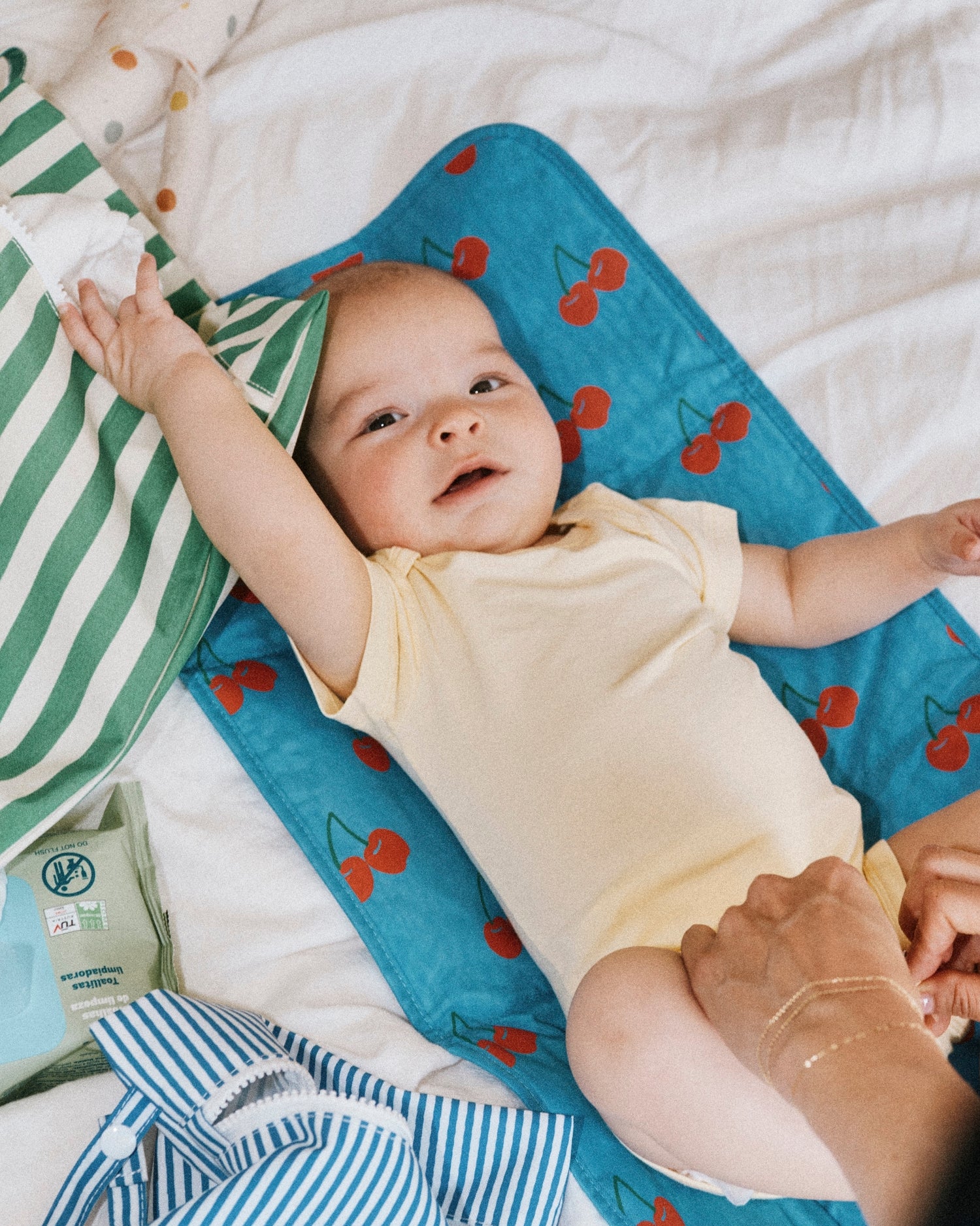 Baby wearing Honey Onesie, from Baby Onesie Two-Pack in Honey Dandelion, laying on Quick Change Kit in Blue Cherries. Next to baby is Wet/Dry Bag Set in Stripe, wet wipes, diapers, doll and white bed background.@model_info:Johnny is wearing 3-6 months.