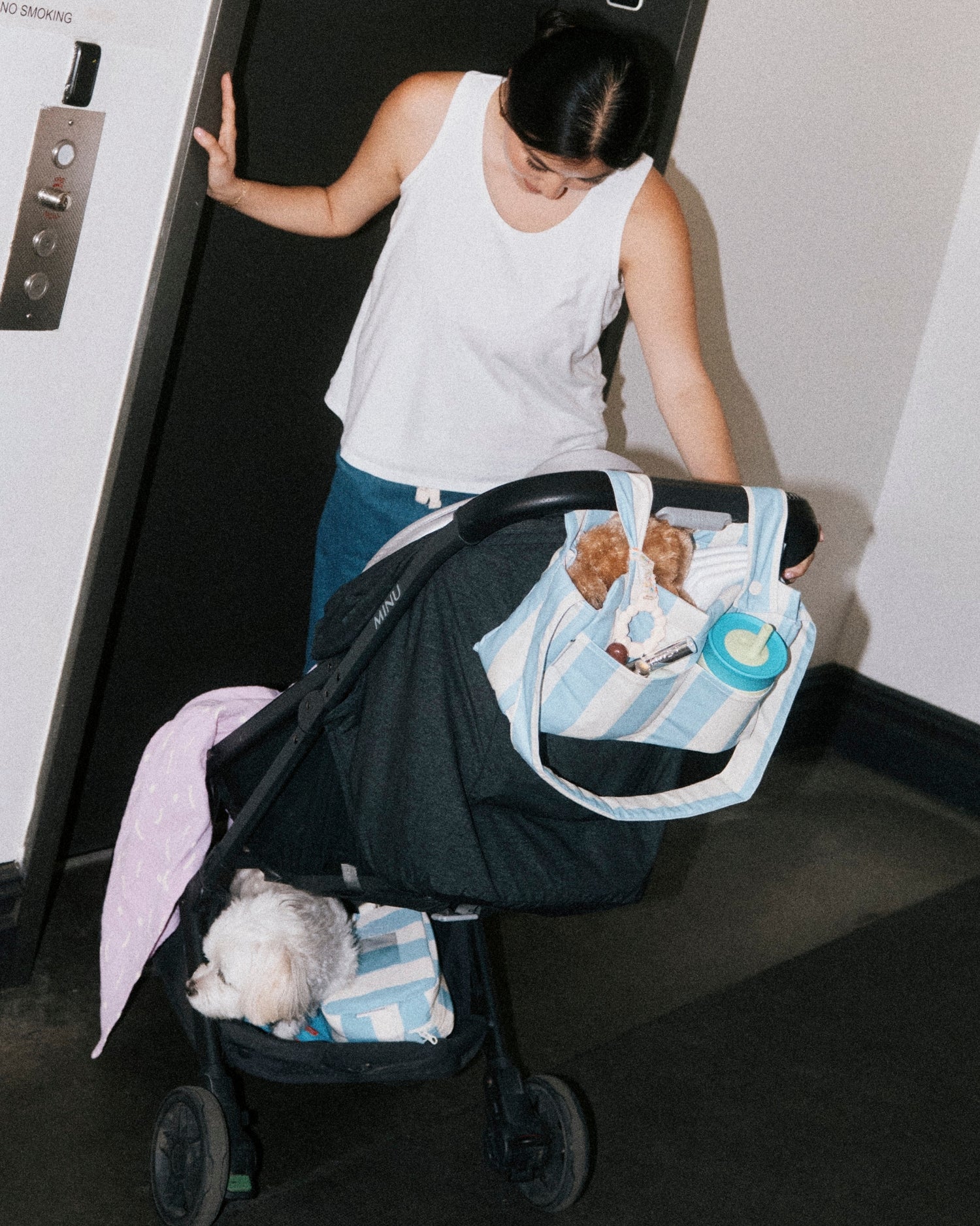 Woman and stroller at elevator, while she looks at baby who is out of view, and covered in Organic Muslin Swaddle in Lilac Squiggles. Stroller holds Stroller Party Caddy Bag in Blue Wide Stripe, full of baby essentials, Mini Cooler Bag in Blue Wide Stripe and a puppy. The woman is wearing Modal Signature Nursing Tank in White. 