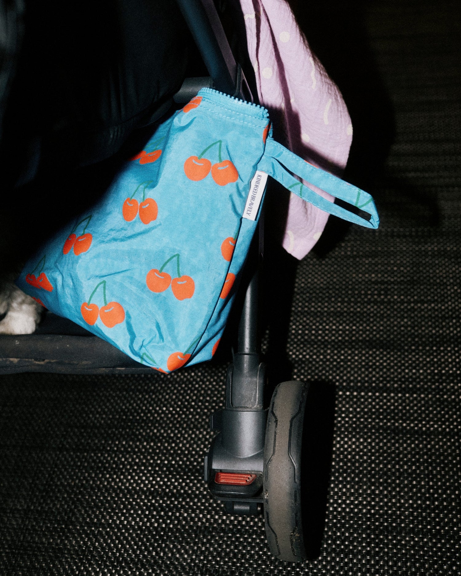 Close up of back side of stroller, in bottom level is Quick Change Kit Bag in Blue Cherries and hanging over side is Organic Muslin Swaddle in Lilac Squiggles.