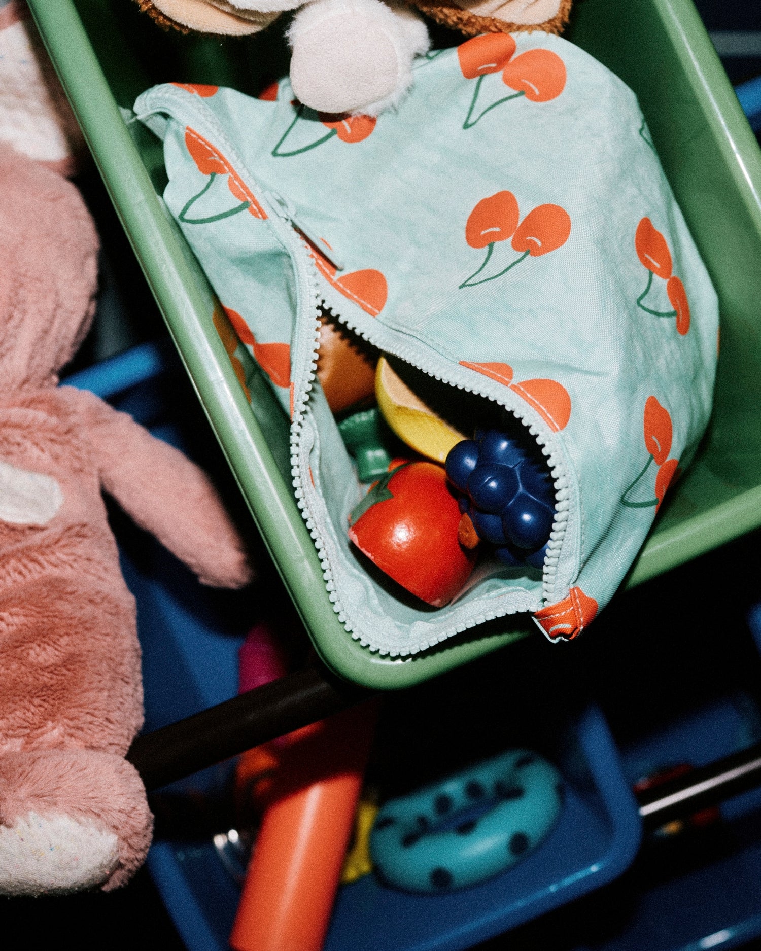 Close up photo of toys in toy home organizer, holding a Zipper Pouch Three-Piece Set in Cherries, and peeking out is a set of wooden fruits and vegetables for baby to play with.