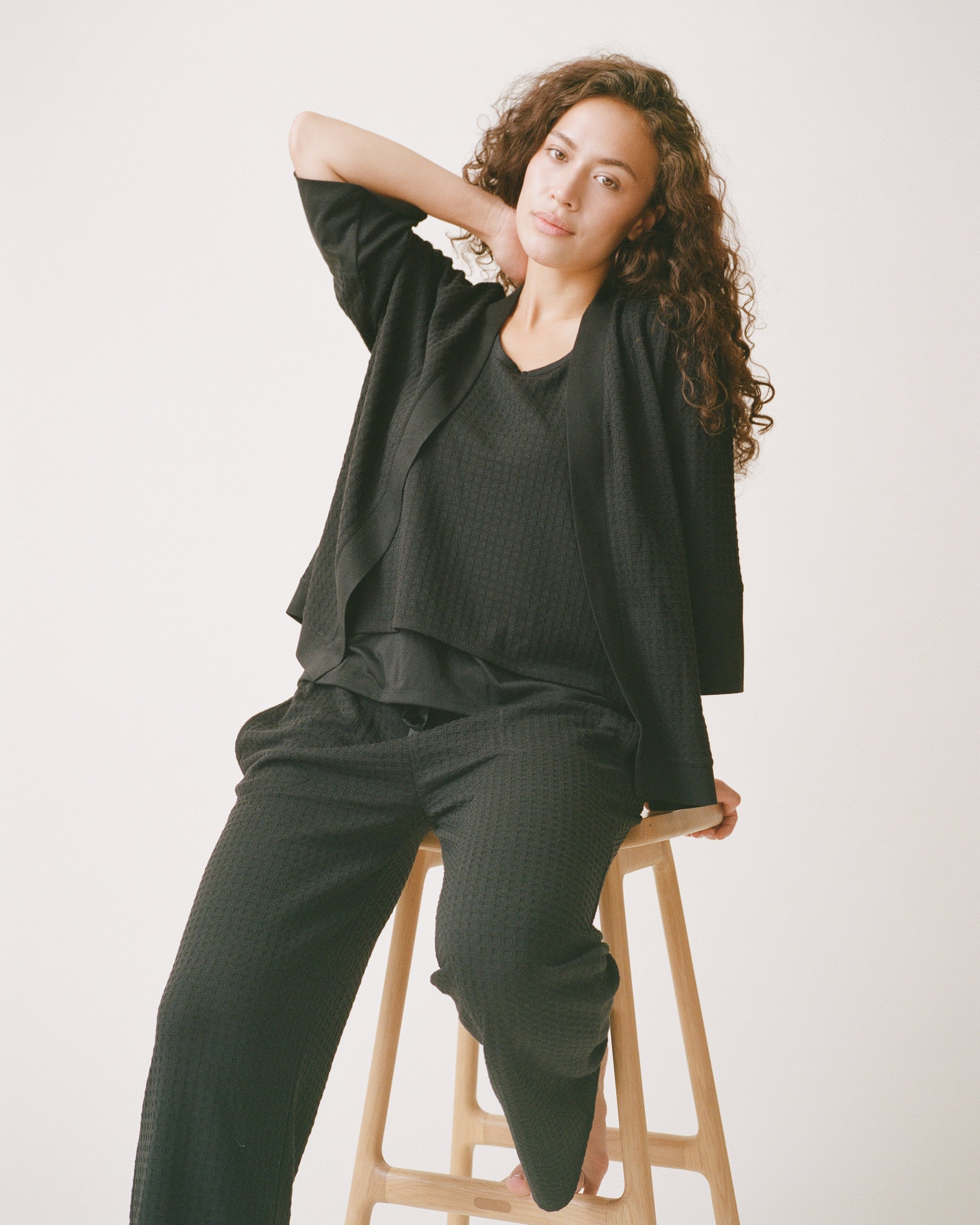 Model is sitting on a stool while wearing Waffle Maternity & Nursing Three-Piece Lounge Set in Black on a beige background.