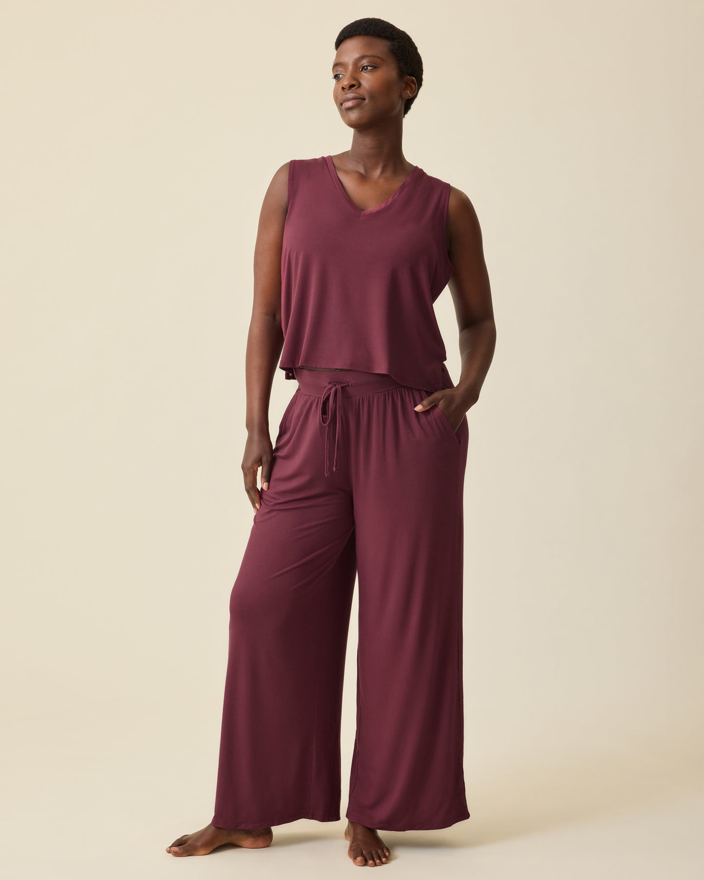 Model is wearing Aria Bamboo Cropped V-neck Tank in Deep Burgundy and Aria Bamboo Casual Lounge Pant in Deep Burgundy on a beige background.