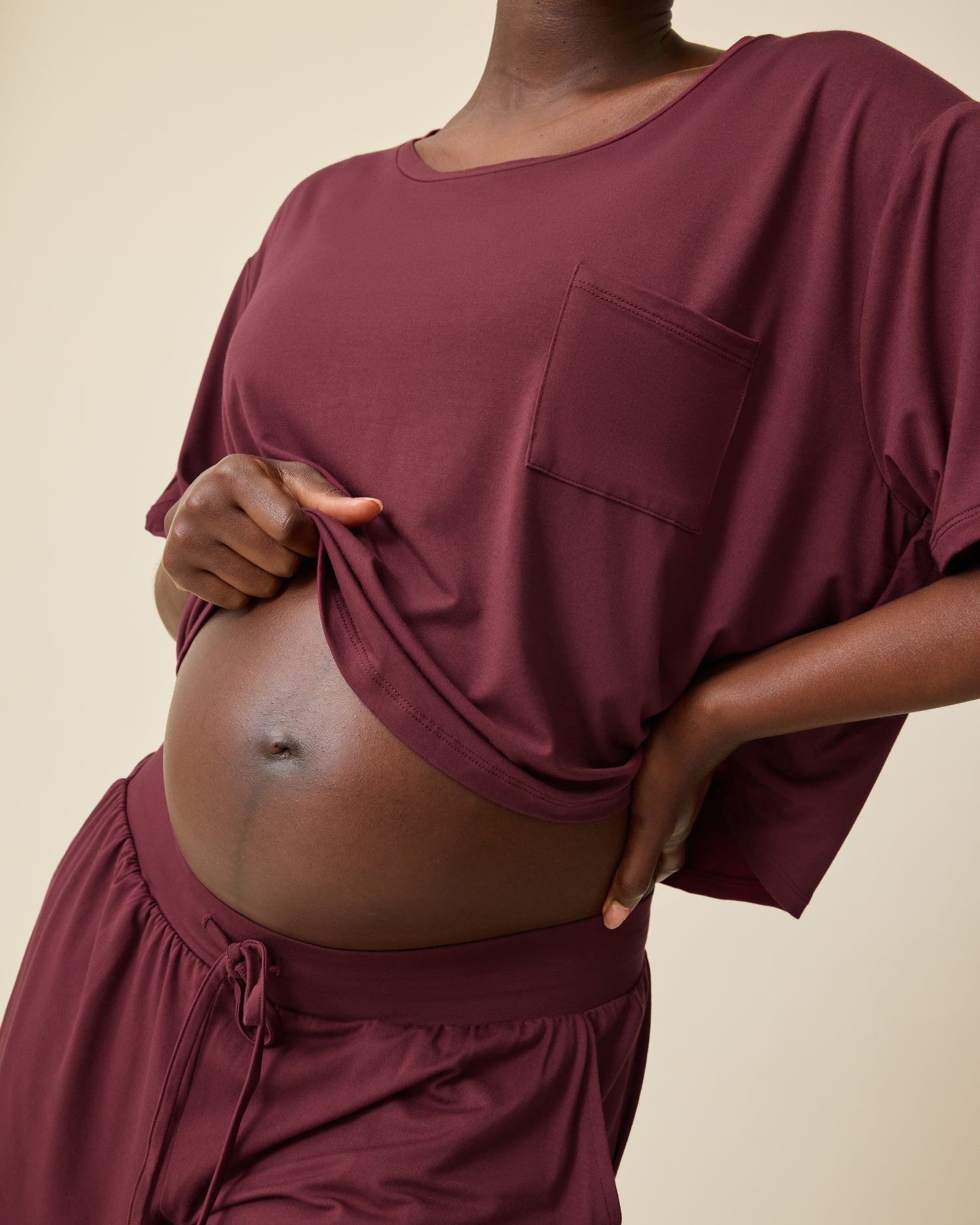 Model is wearing Aria Bamboo Casual Cropped Short Sleeve Top in Deep Burgundy, pulling up top to show baby bump, and Aria Bamboo Casual Lounge Short in Deep Burgundy, on a beige background. 