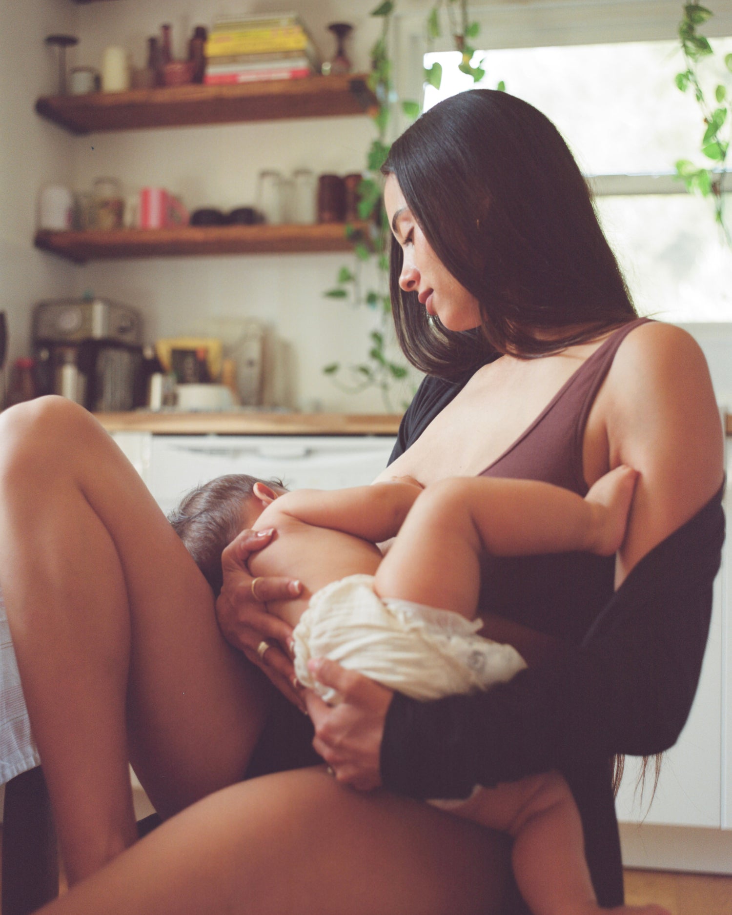 Mother breastfeeding newborn while wearing Every Stage Crossover Bra in Cocoa in a home kitchen setting