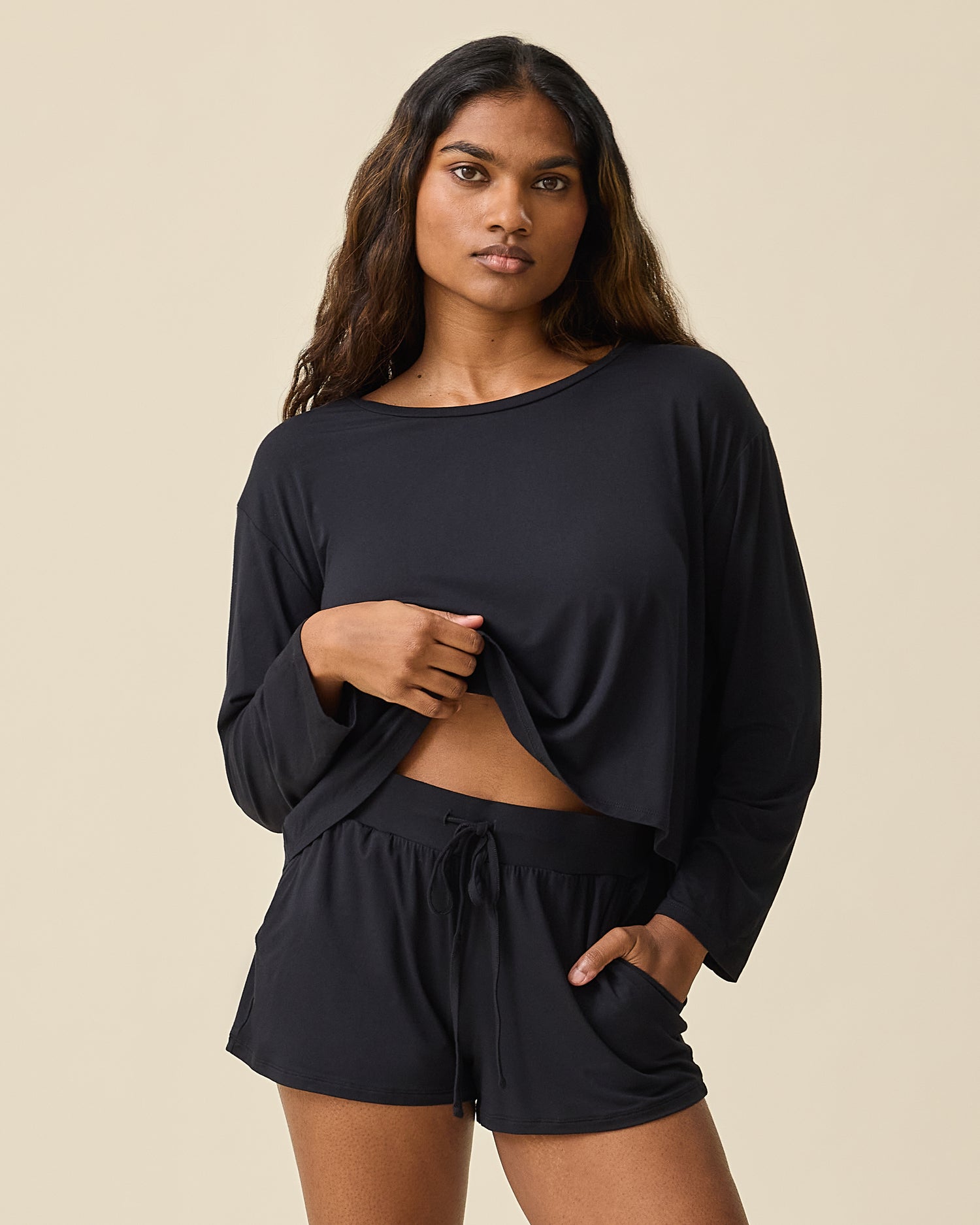 Woman wearing the Aria Bamboo Casual Lounge Short in black with matching long sleeve top lifting top to show waistband and drawstring on a beige background