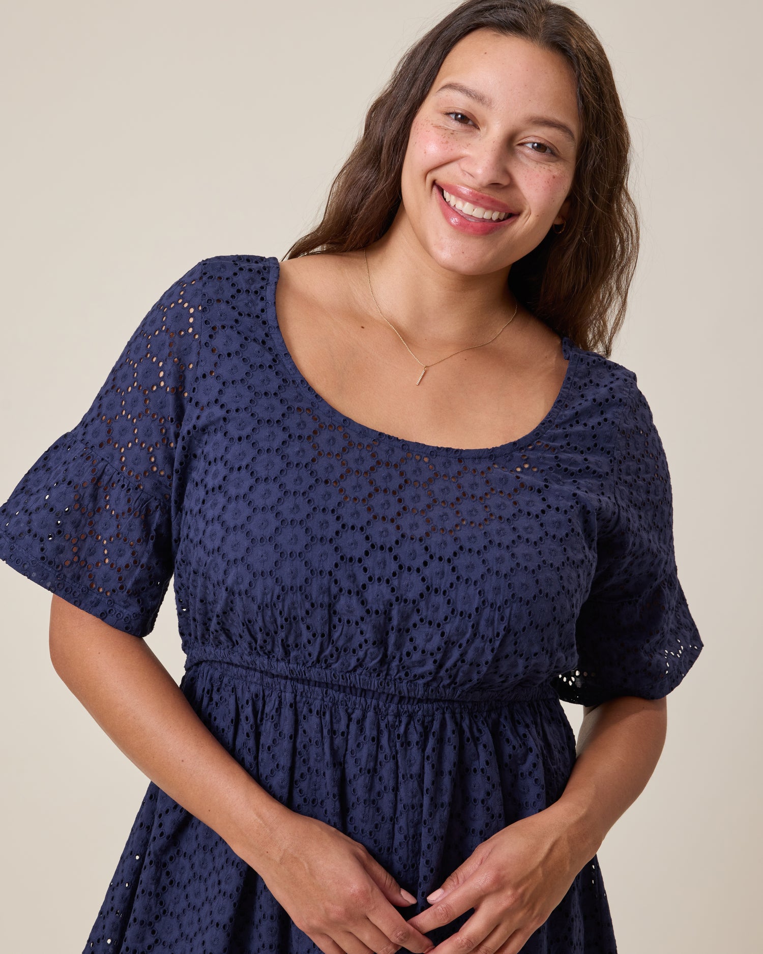 Woman wearing the Dahlia Eyelet Maternity & Nursing Dress in Navy with a textured pattern against a beige background