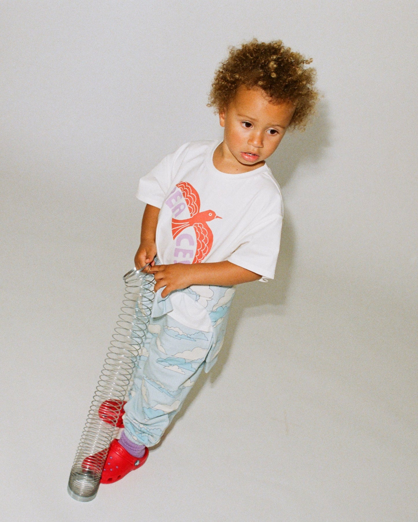 Model is wearing Graphic Tee in White Bird and French Terry Jogger in Blue Clouds while playing with a slinky, on a gray background.