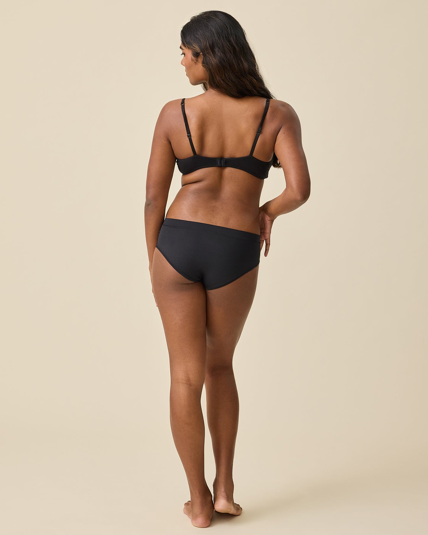 Back view of model wearing Grow with Me™ Maternity & Postpartum Hipster in Black on a beige background