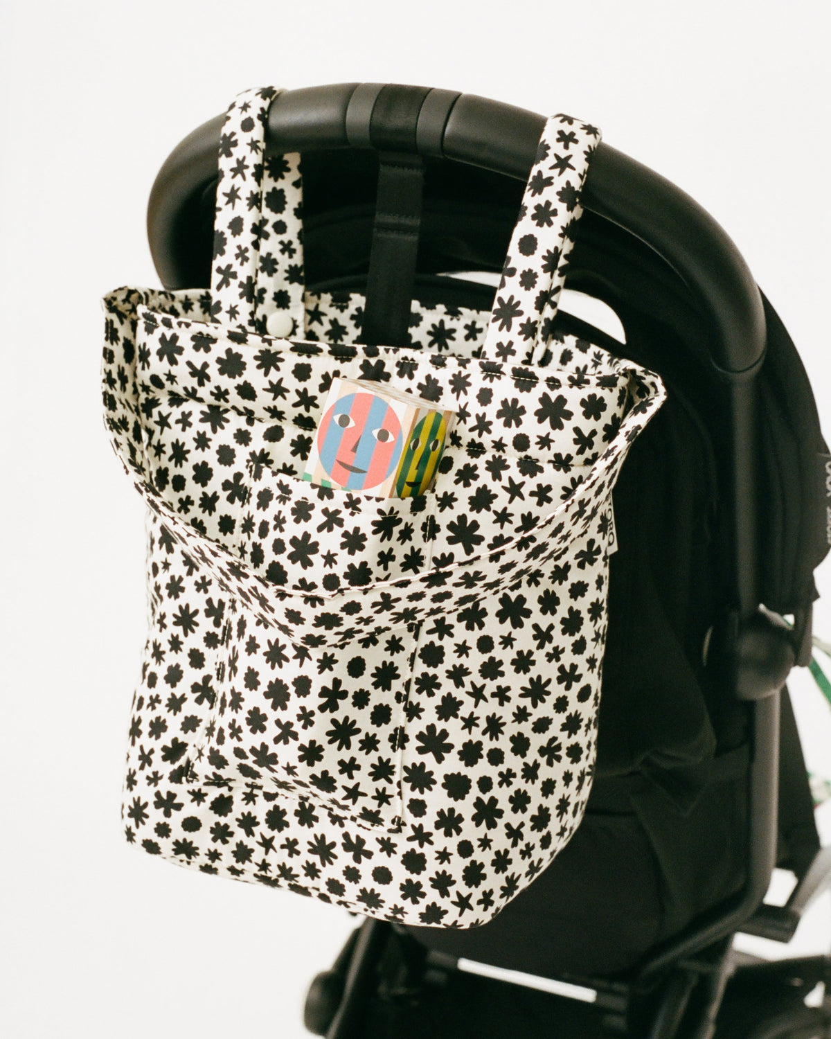 Stroller Party™ Bucket Bag in Cream Chamomiles is hanging off the handlebar of a black stroller with wooden whistle toy peeking out of pocket, on a beige background.