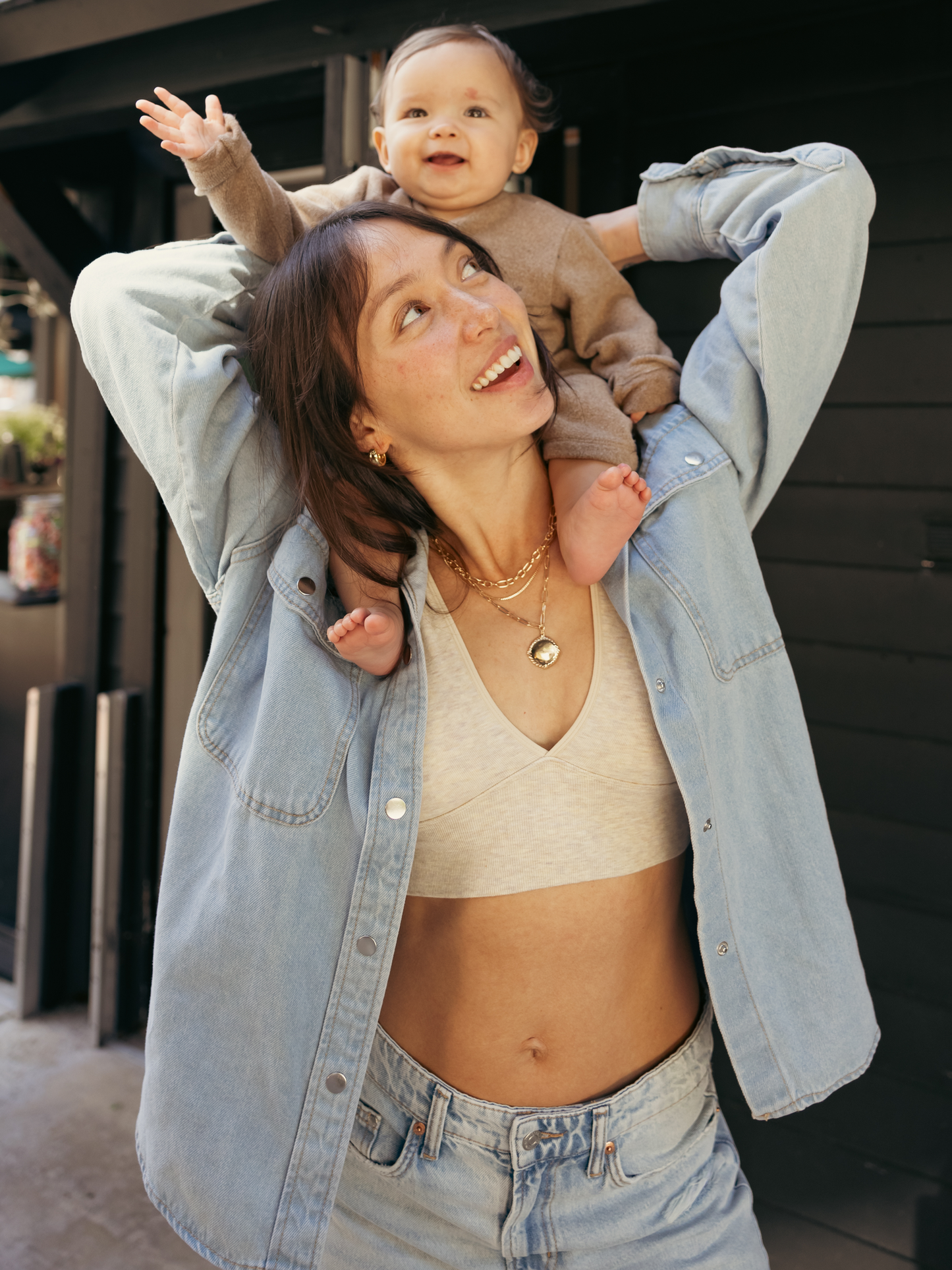 Model wearing the Plunge Neckline Maternity & Nursing Bra in Oatmeal Heather styled with a denim shirt. Carrying her baby on her shoulders. 