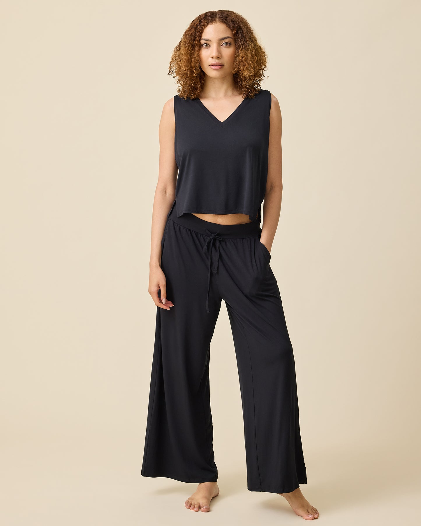 Model is wearing Aria Bamboo Cropped V-neck Tank in Black and Aria Bamboo Casual Lounge Pant in Black on a beige background.