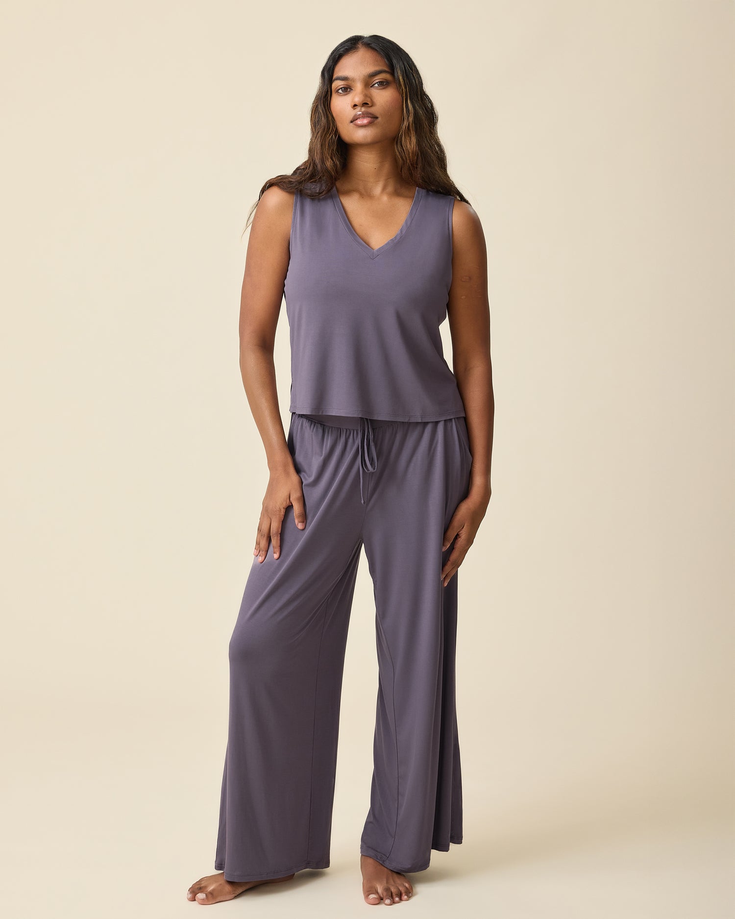 Model is wearing Aria Bamboo Cropped V-neck Tank in Granite and Aria Bamboo Casual Lounge Pant in Granite on a beige background.